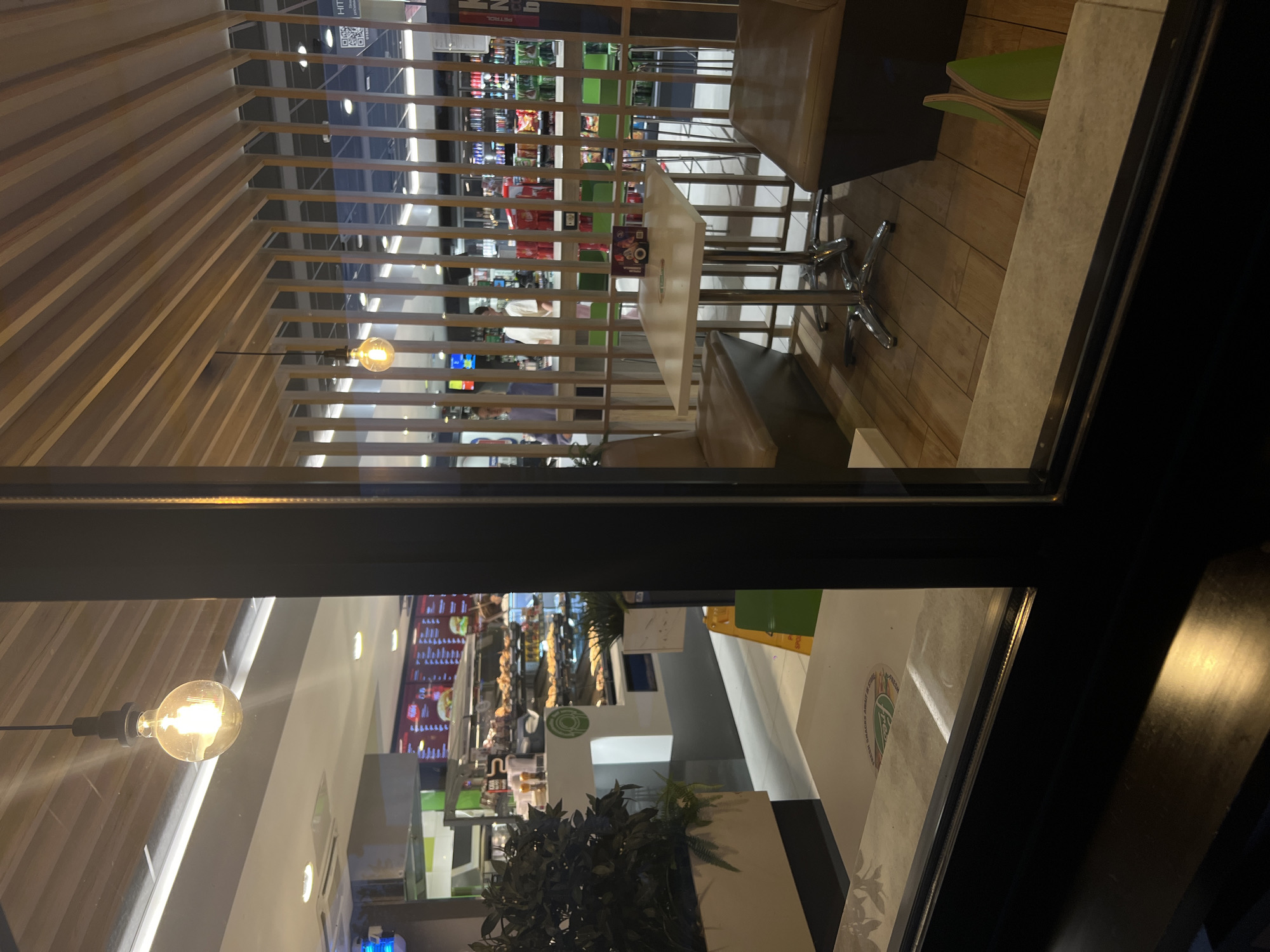 The image shows an interior view of a McDonald's restaurant in Šempeter pri Gorici, Slovenia. The scene is viewed from outside, through a window, at night. Inside, a seating area with light brown booth seats and a light-colored square table with a chrome base is visible. The table has a small, colorful box on it. Beyond the seating area, a serving counter with food displays and menus is visible. Several people are working behind the counter, and customers appear to be present. The interior features a modern design with wooden slatted ceiling and partitions. A hanging pendant light with a visible bulb illuminates the foreground seating area. The floor in the seating area is wooden planking. In the background, shelves stocked with various products are visible.