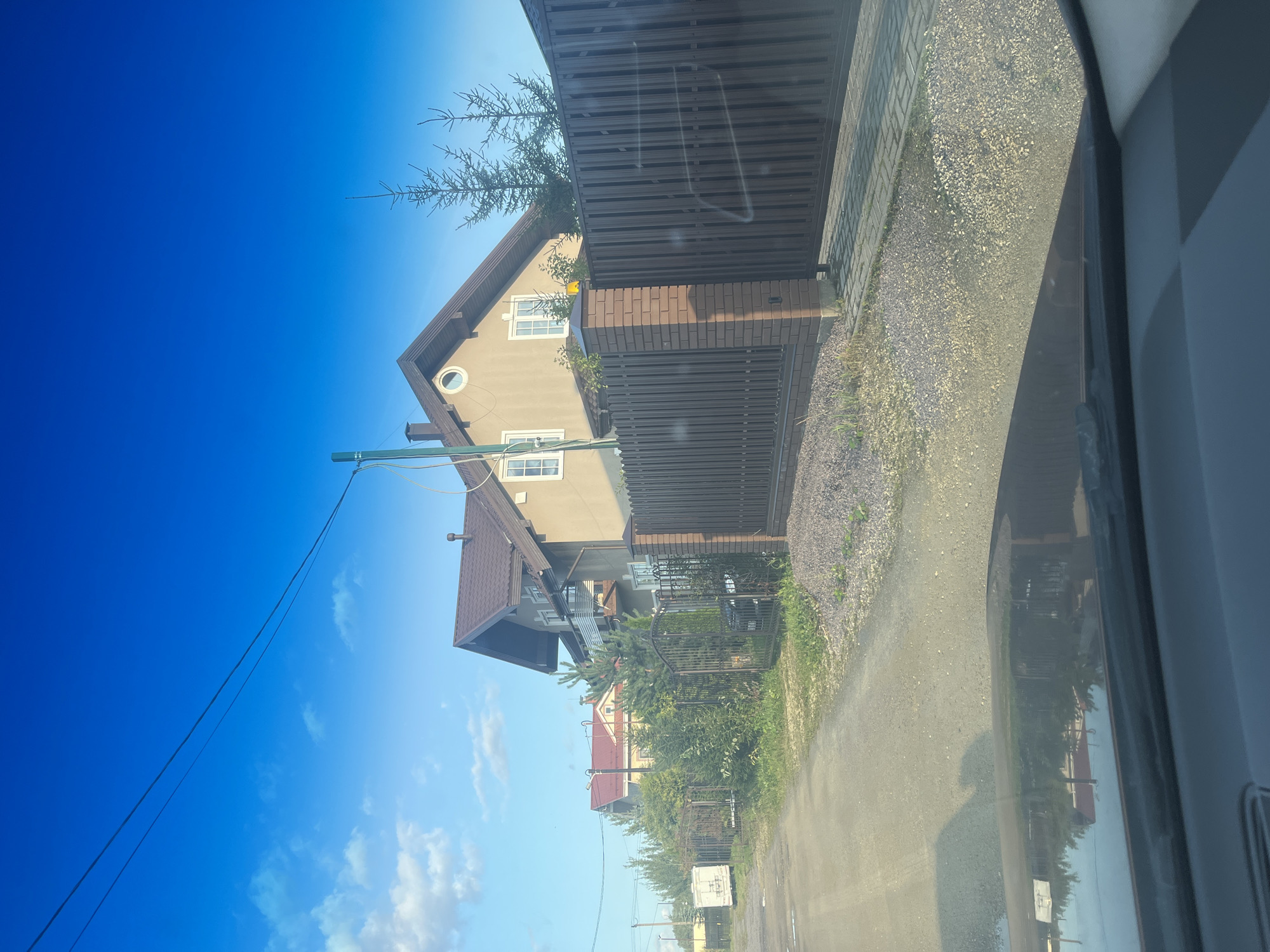 The image depicts a residential street in Vladimirovka, Russia, on a clear, sunny day. The view is from inside a vehicle, looking out through the windshield. Two houses are visible, one in the foreground with a beige facade, dark brown metal fencing, and a brick gatepost, and another house further back with a red roof. Evergreen trees and dense foliage line the street. A white utility trailer is parked on the left side of the road. The sky is a vibrant blue with scattered white clouds. The road surface appears to be a mix of gravel and dirt. The reflection of the houses and trees is visible on the vehicle's windshield.