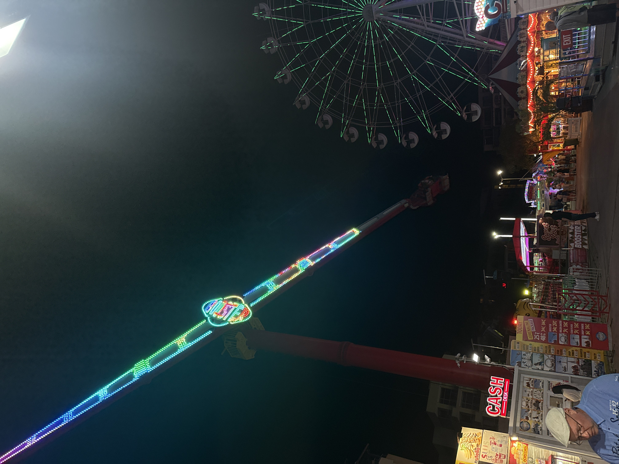 The image captures a nighttime scene at an amusement park, likely located in Nesebar, Bulgaria, given the context. A prominent thrill ride, a pendulum-style swing ride illuminated with colorful LED lights, dominates the left side of the frame. Its long arm extends diagonally upwards against the dark sky, with a gondola at its end. To the right, a Ferris wheel, also outlined in green lights, stands tall, its cabins visible.
In the foreground and midground, various stalls and attractions are set up. A "CASH DESK" sign glows red above a booth displaying a menu of rides and prices, suggesting a ticket sales or information point. People are milling about, though they are not the primary focus of the image. Some are standing near the booths, while others appear to be walking through the park. The overall atmosphere is typical of a carnival or fairground at night, with artificial lights illuminating the rides and structures. Buildings can be seen in the background, indicating an urban or semi-urban setting. The ground appears to be paved, consistent with a public walkway.