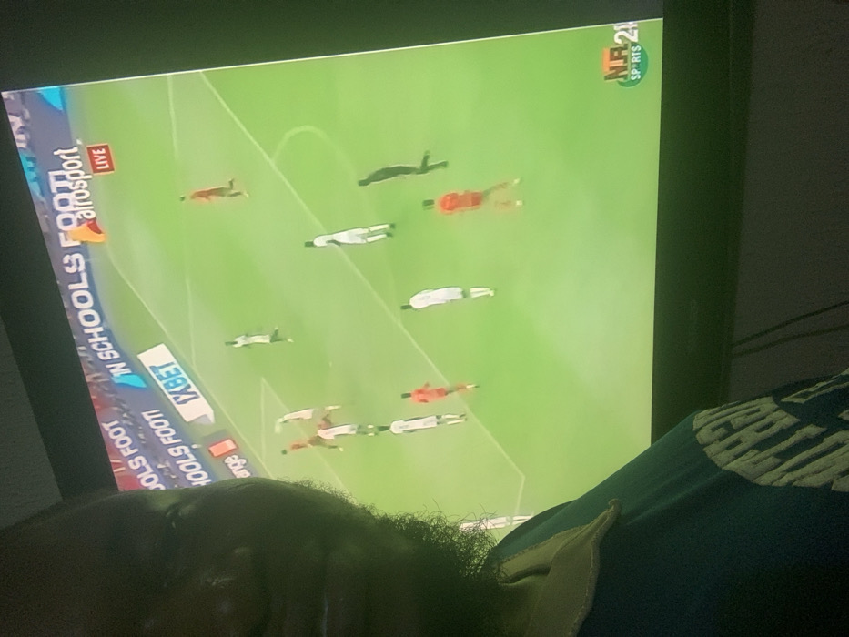 A person is watching a soccer game on a television. The screen displays a top-down view of a field with players in red and white uniforms, and their shadows are visible. The broadcast includes logos for "N SCHOOLS FOOTBALL", "alrosport LIVE", and "RTS". The person appears to be lying down, possibly sleeping. Part of their hair and t-shirt is visible in the foreground.