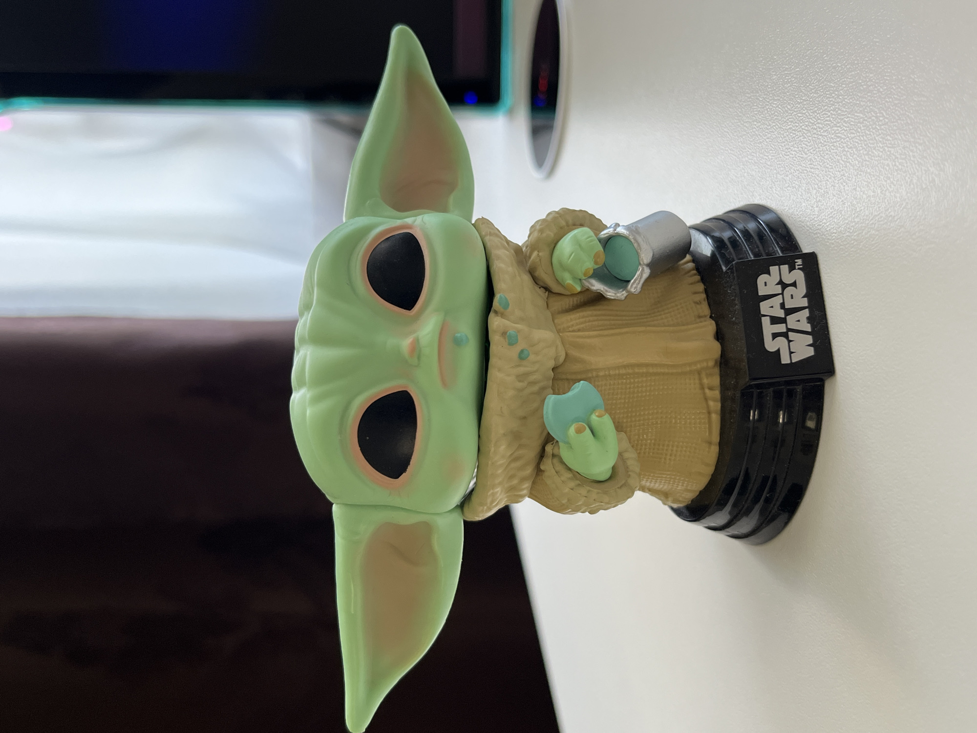 A Funko Pop figure of Grogu from Star Wars is positioned on a white, textured surface. The figure is predominantly green with large ears, black eyes with pink accents, and is wearing a tan robe with fur trim. Grogu is holding a light blue sphere in its left hand and a silver cup with a blue sphere inside its right hand. The figure stands on a black base with "STAR WARS" branding. The background is blurred, showing dark brown curtains on the left and a white curtain and a dark monitor with blue and green accents on the right. This image was captured in Babruysk, Belarus.