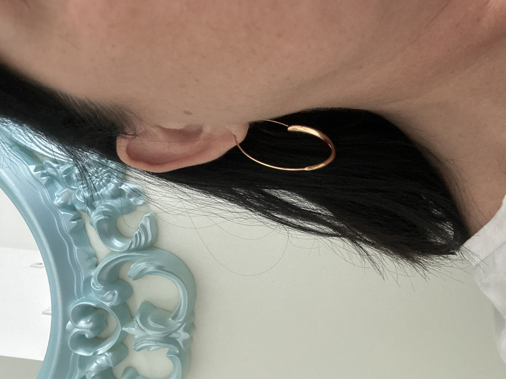 A close-up shot focuses on a person's ear adorned with a gold hoop earring. The ear is pink, and the hoop is positioned so the closure is near the top of the ear lobe. Dark black hair partially obscures the ear, cascading towards the bottom right of the image. To the left, a fragment of a light blue ornate frame, possibly a mirror, is visible. The background appears to be a plain, light-colored wall. The image is taken from an angled perspective, looking slightly upward. There is no indication of this being in Saint Petersburg, Russia.