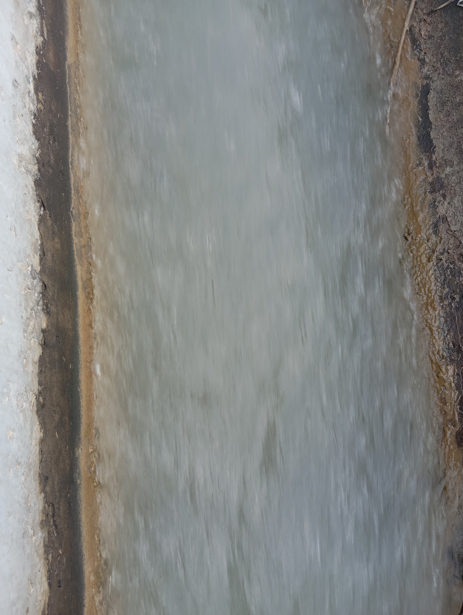 The image shows a close-up, blurred view of a narrow channel of water flowing over a rocky, earthen bank. On the left side, there is a light-colored, rough surface that appears to be stone or concrete, with a dark, irregular line separating it from the water. The water itself is a pale, milky gray and appears to be in motion, creating streaks and blurs. The right bank of the channel is rough and textured, with shades of brown and dark gray, and a thin, dry twig is visible lying on it. The overall impression is of a man-made or natural water channel, possibly in a rural or undeveloped area. The specific location within Kızıl-Bel, Kyrgyzstan, cannot be determined from the image alone.