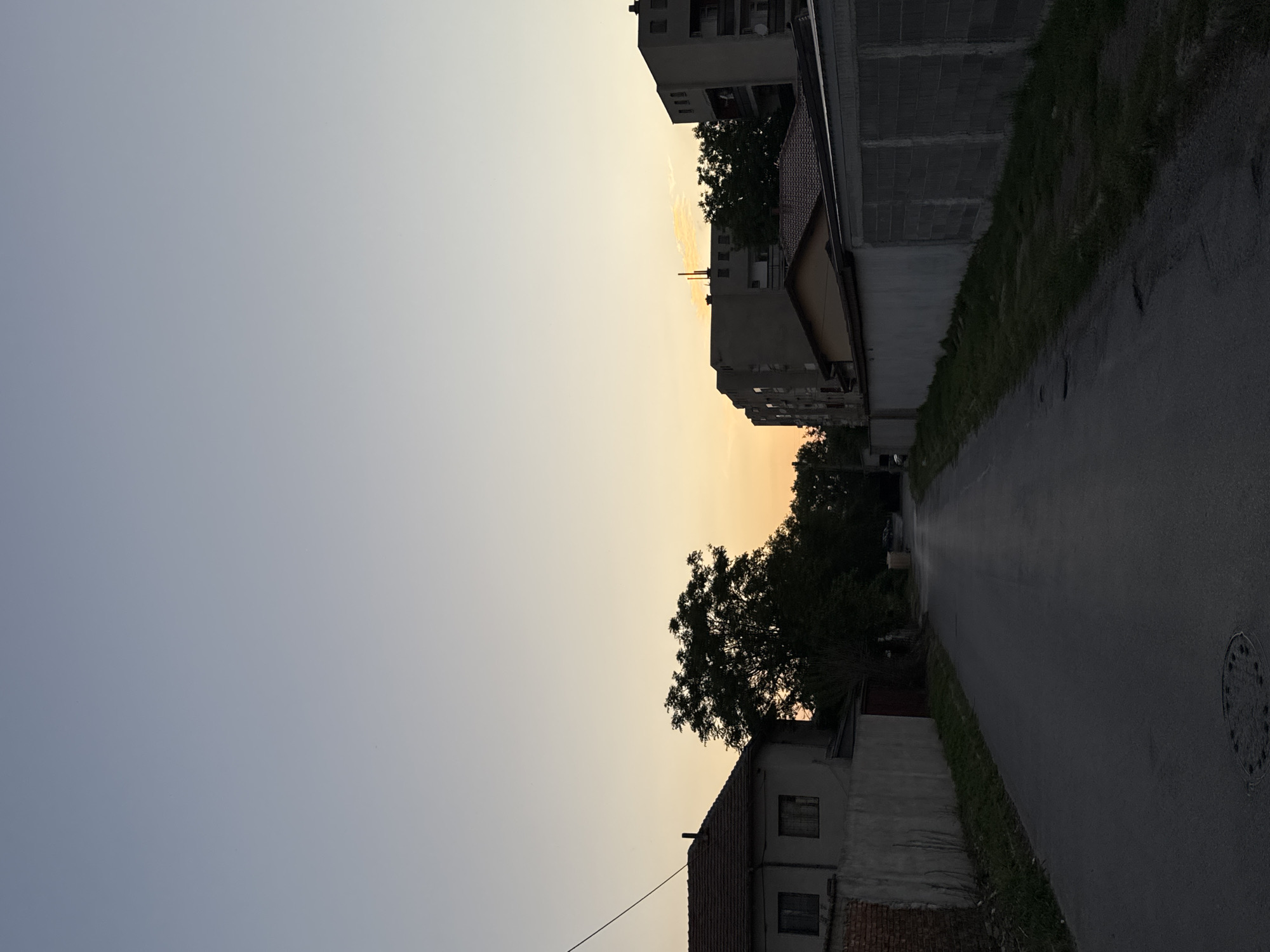 The image depicts an urban street scene at dusk in Unknown, Unknown. The sky is a gradient of light blue to orange, indicating the setting sun. On the left side, a few residential buildings are visible. The road curves to the right, bordered by concrete walls and green grass. A few trees are visible, silhouetted against the sky. A manhole cover is visible in the road on the right. No people are visible in the image.