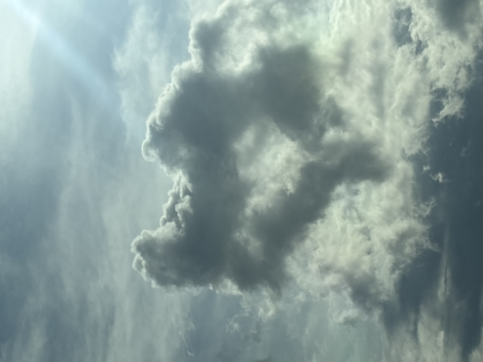 The image captures a cloud formation in the sky over Сеніцкі сельскі Савет, Belarus. The clouds are predominantly dark gray with lighter highlights, suggesting varying densities and possibly approaching weather. The overall lighting indicates it is daytime, with a faint sunbeam visible in the upper right quadrant. No people, structures, or discernible geographical features are visible in this close-up of the sky.