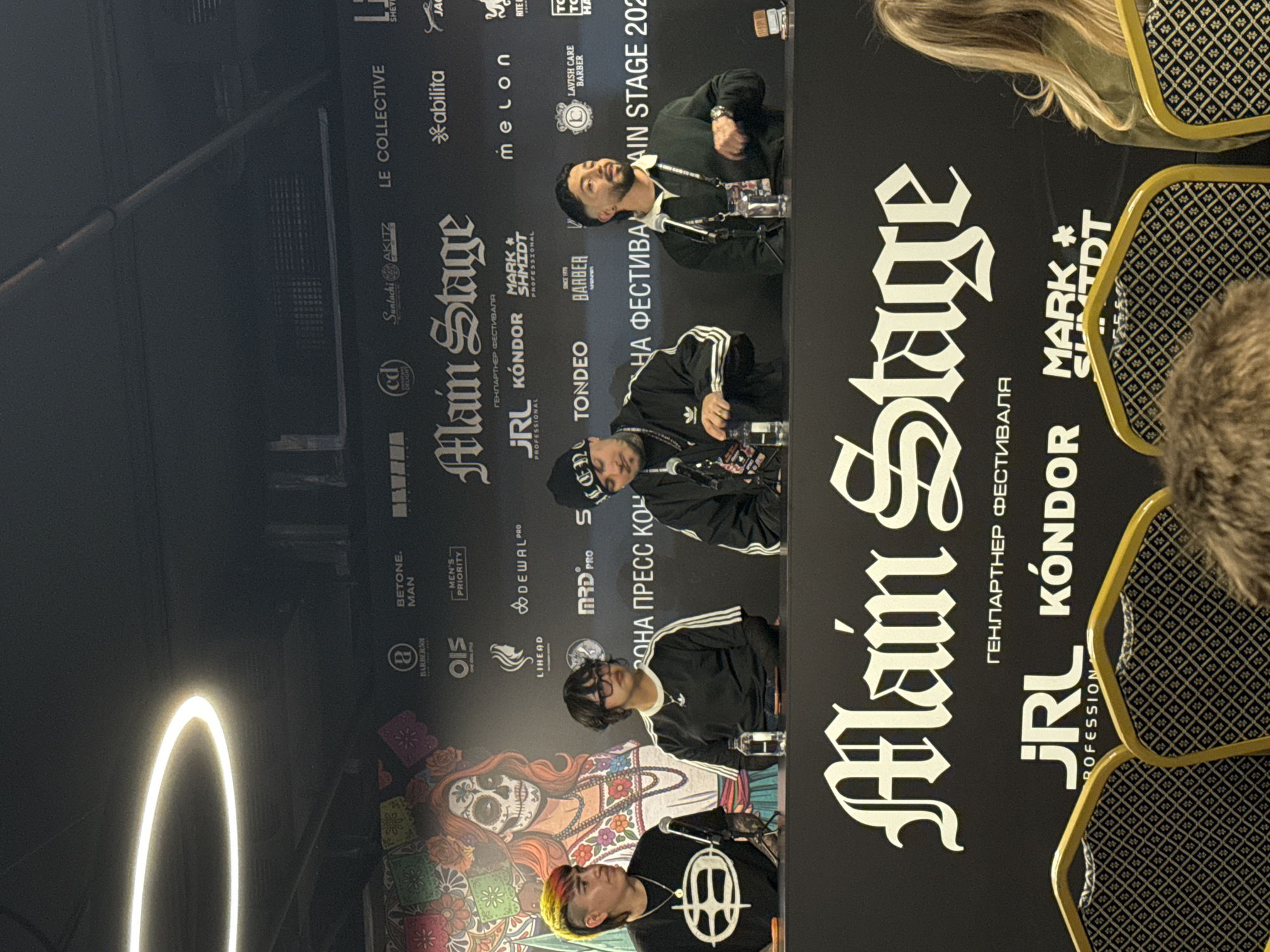 A medium shot of a press conference backdrop, likely from the Main Stage festival based on the prominent logo. Four individuals are seated at a table facing the camera. The backdrop features logos of various sponsors, including JRL Kondor, Mark * Shmidt Professional, and others. The backdrop is predominantly black with silver and white fonts, showcasing a Day of the Dead-style illustration at the bottom left. The environment seems to be indoors, and the location (Khimki, Russia) is inferred from the Russian text on the backdrop. The four individuals are dressed in casual attire.