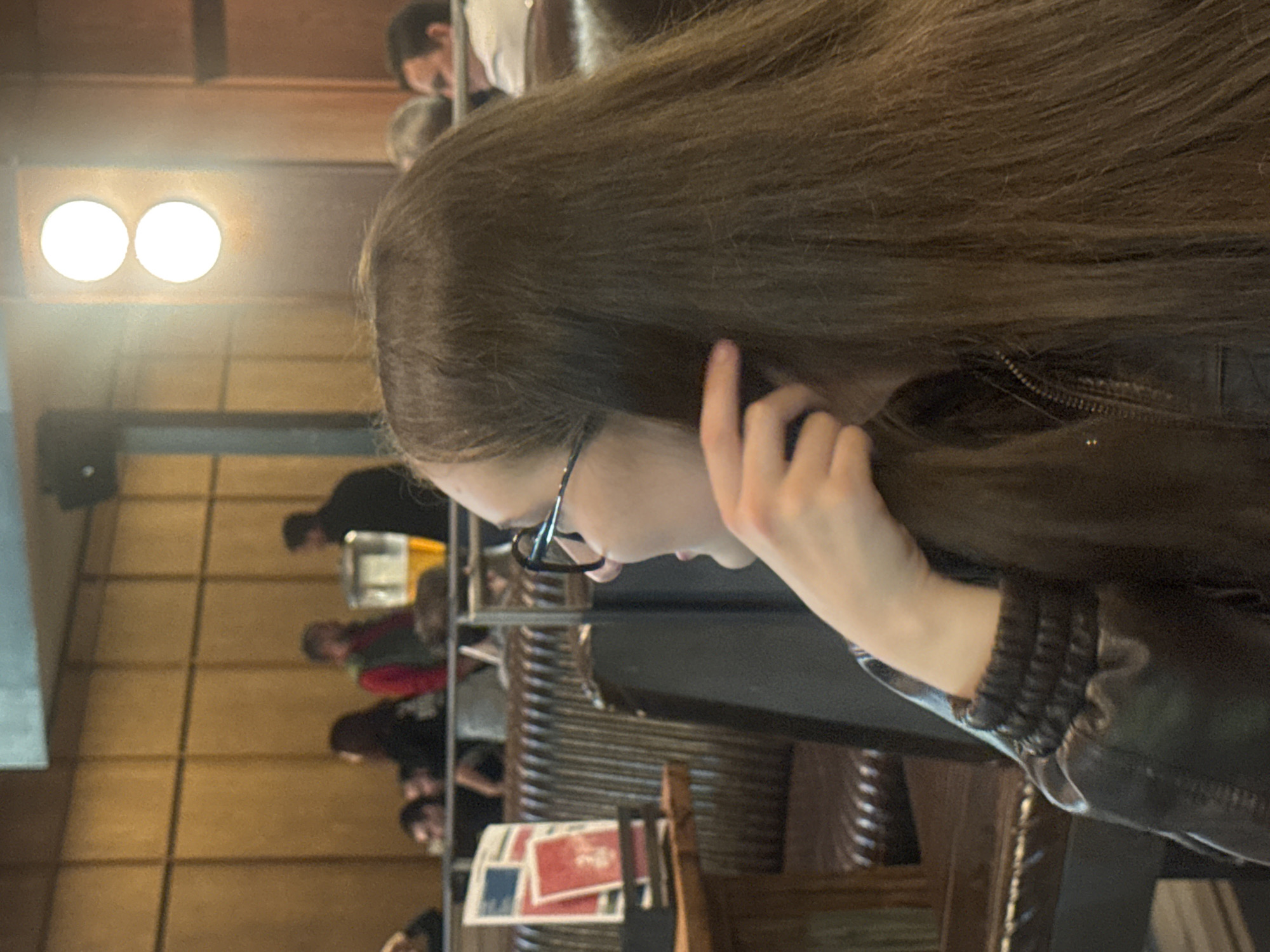 A woman with long brown hair and glasses is seated, wearing a black jacket with a ribbed cuff. She is touching her hair near her ear. The setting appears to be a restaurant or cafe interior with wood paneling and chairs. In the background, other people are seated at tables. There are menus or papers on the table in front of the woman. The location is in Tbilisi, Georgia.