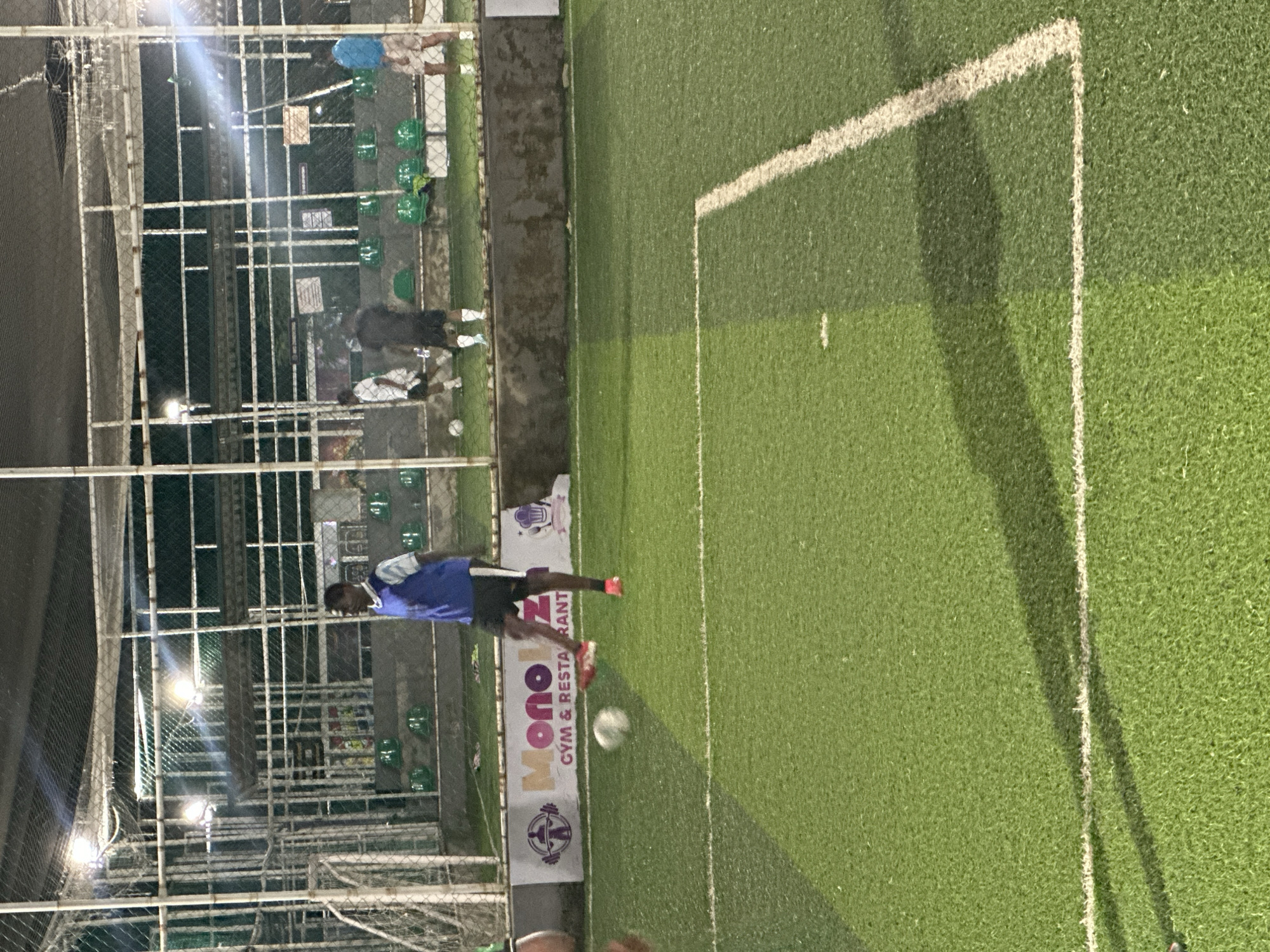 The image shows an outdoor artificial turf soccer field at night in Abuja, Nigeria. A man wearing a blue vest over a white and blue striped shirt and black shorts is playing soccer. He is in mid-action, with a soccer ball in the air in front of him, suggesting he is about to kick it. In the background, other individuals are visible on the field, and there are green stadium seats and a fence. A banner with "MonoGym & Restaurant" is displayed along the edge of the field. The lighting suggests it is an indoor or enclosed outdoor facility, possibly a training center or recreational complex.