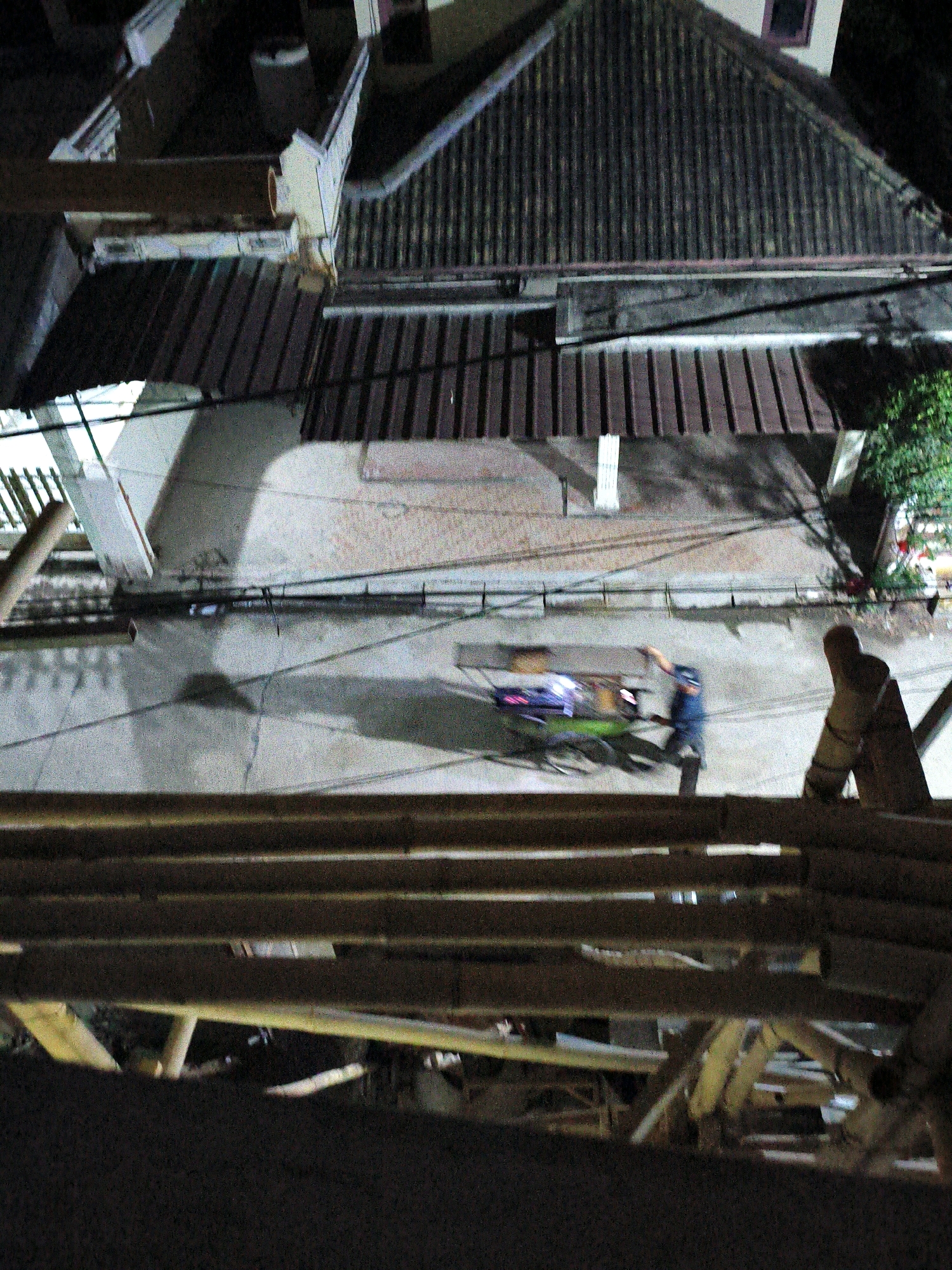 In an overhead, grainy night-time shot from an elevated perspective, a scene unfolds in a street in Bekasi, Indonesia. The foreground is dominated by a blurry, textured wooden structure, possibly scaffolding or a fence, obscuring a portion of the view. Below this, the dark asphalt street is visible, illuminated by ambient light casting long shadows.
Two individuals are present on the street. One, wearing a dark blue top and pants, is actively pushing or maneuvering a cart. The cart itself is illuminated with bright, colorful lights, suggesting it is a food cart or mobile vendor. The second individual, possibly associated with the cart, is partially obscured and in motion, their form blurred by the camera's low light conditions.
The background reveals buildings with pitched, dark-colored roofs, typical of residential or commercial structures in an urban environment. The lighting suggests it is nighttime. The overall impression is a glimpse of everyday street life in Bekasi after dark, captured with limited clarity due to the photographic conditions.