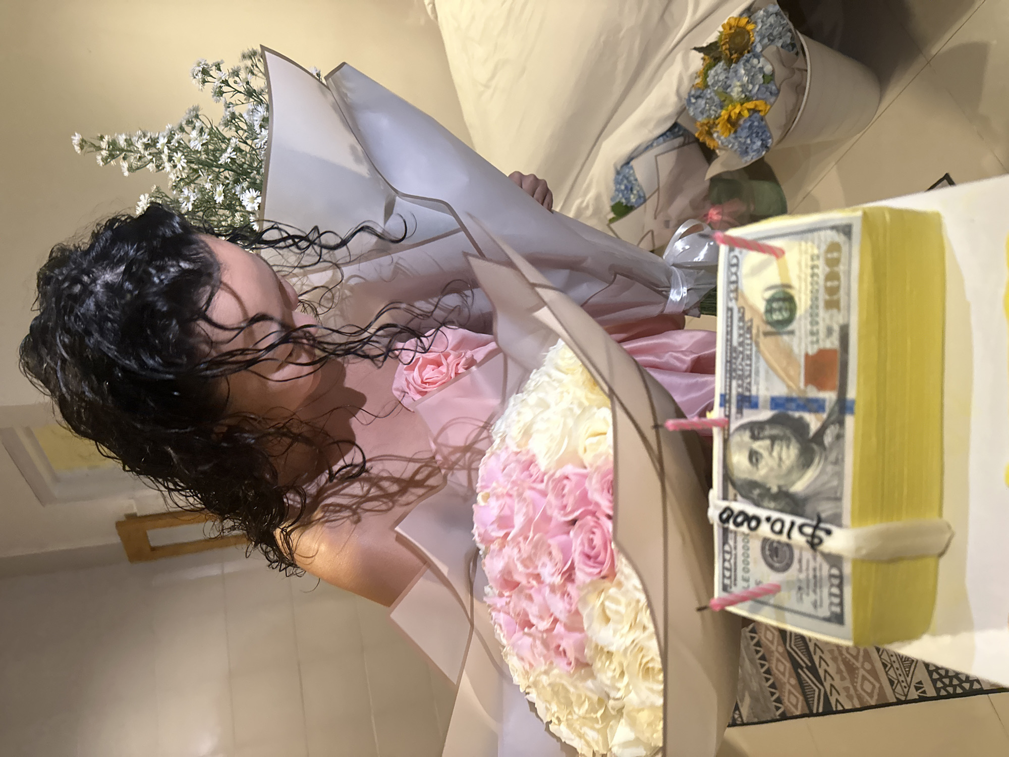In a well-lit room, possibly a hotel or apartment in North Kuta, Indonesia, a young woman with long, dark, wet hair is holding a bouquet of white and pink roses. She is wearing a pink satin garment. In front of her is a cake decorated to resemble a stack of hundred-dollar bills, complete with two lit pink candles and a handwritten tag that reads "$10.000". To her right, another bouquet of small white flowers is visible, and further to the right, a small bucket holds a cluster of blue hydrangeas and yellow flowers. The background consists of a wall and a bed with white linens.