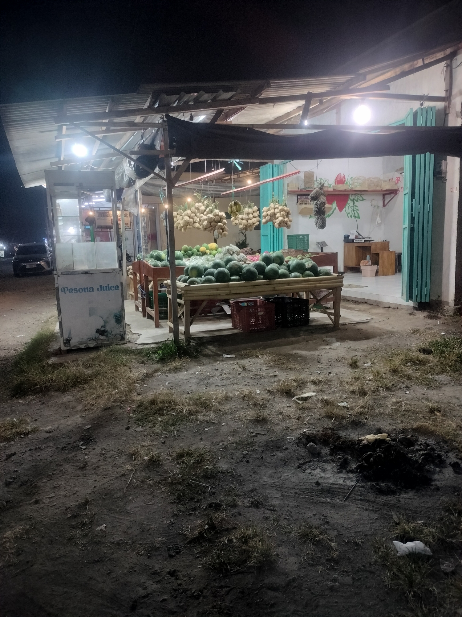 This image depicts an outdoor fruit stand at night in Srono, Indonesia. The stand is constructed of wood and corrugated metal, with a tarp providing overhead cover. Fruits, including watermelons and what appear to be onions or garlic bulbs, are displayed on wooden tables. A "Pesona Juice" sign is visible on a white kiosk. In the background, a building with light blue folding doors is visible, along with some interior furnishings. A dark-colored car is parked on the left side of the image. The ground in front of the stand is unpaved and appears to be dirt with sparse vegetation and some debris, including a small pile of ash and burnt material. The lighting suggests artificial illumination from the stand itself and possibly from the building in the background.
