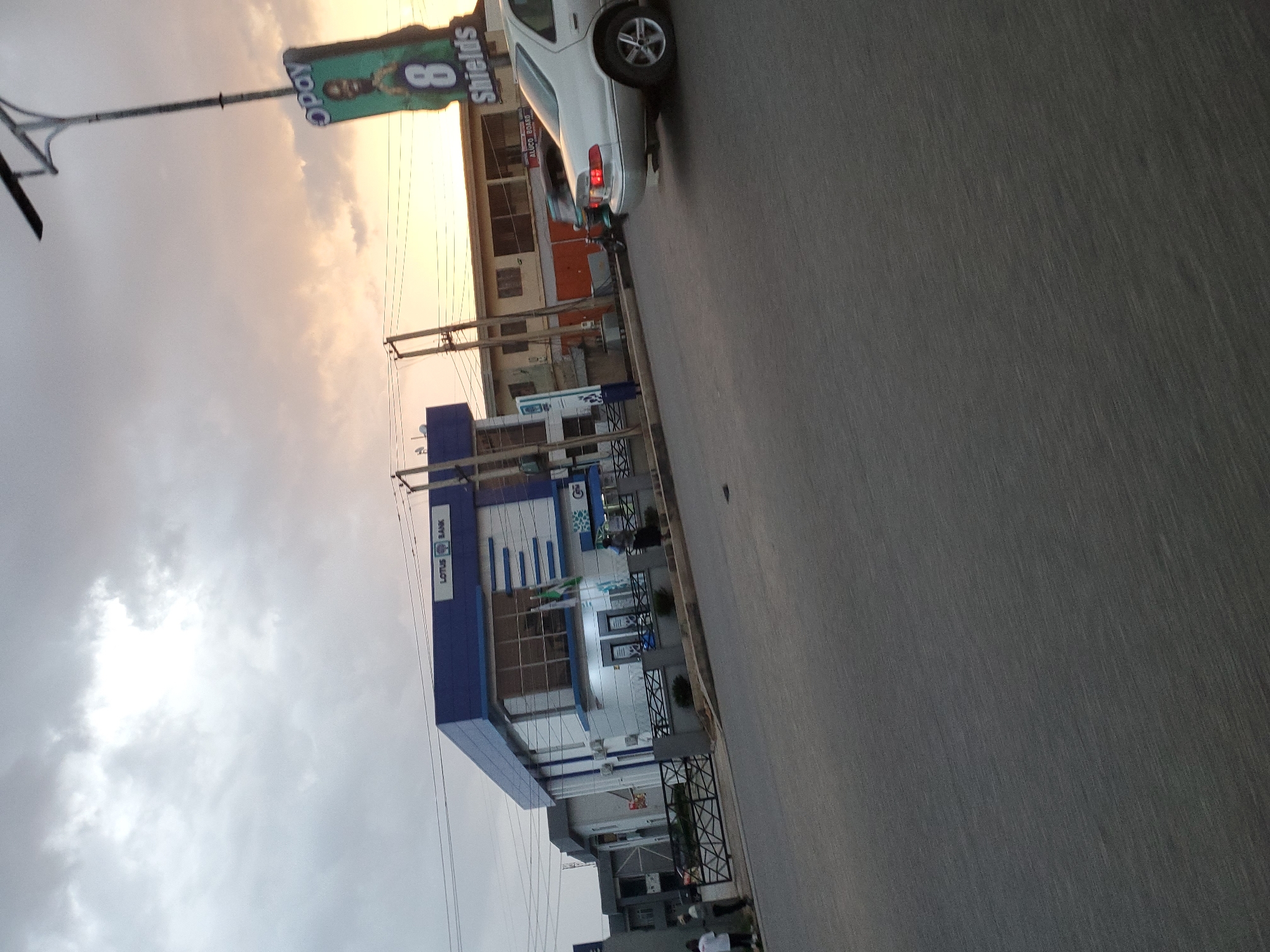 The image depicts a street scene in Jos, Nigeria, with a building labeled "LOTUS BANK" prominently featured. The sky is overcast with hints of sunset colors. In the foreground, a silver sedan is parked on the right side of the road. A billboard for "OPay 8 Shields" is visible on a lamppost. Several people are seen near the bank and on the sidewalk, and a motorcycle is in motion on the street. The overall environment suggests an urban setting.