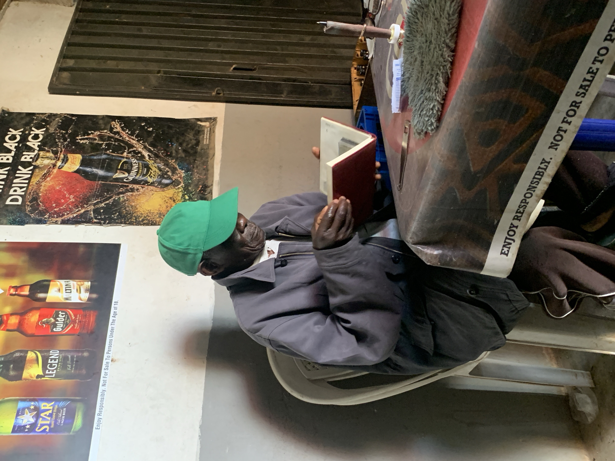 A man wearing a green baseball cap and a gray jacket sits at a table in a room in Barakin Lerawi, Nigeria. The man is looking down at an open red book in his hands, and appears to be reading. The table is covered with a red and brown patterned cloth, and there are various items on the table, including a small metal object, a dust brush, and a small white object. Behind the man, there is a poster on the wall advertising Guinness. The room has a concrete wall and a metal door. The lighting is dim.