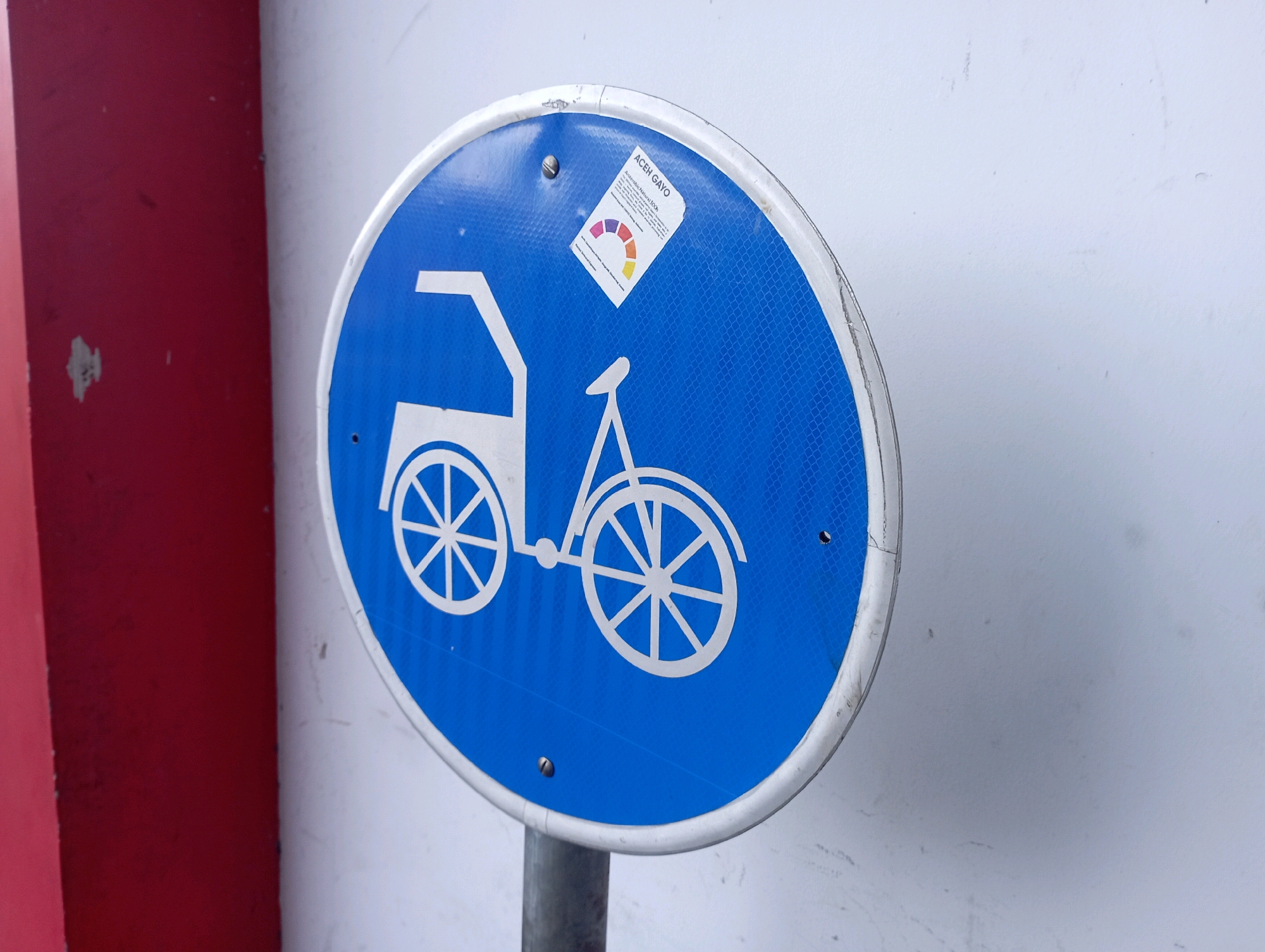 The image shows a blue circular road sign with a white pictogram of a bicycle and a person in a carrier. A small sticker is attached to the upper right portion of the sign. The sign is mounted on a metal pole. To the left of the sign, a red wall is visible. The background is a white wall. The scene appears to be outdoors, possibly on a street or sidewalk in the city of Ponorogo, Indonesia, based on the sticker text "ACEH GAYO" which may indicate a regional connection or product. The lighting suggests it is daytime. There are no people visible in the image.