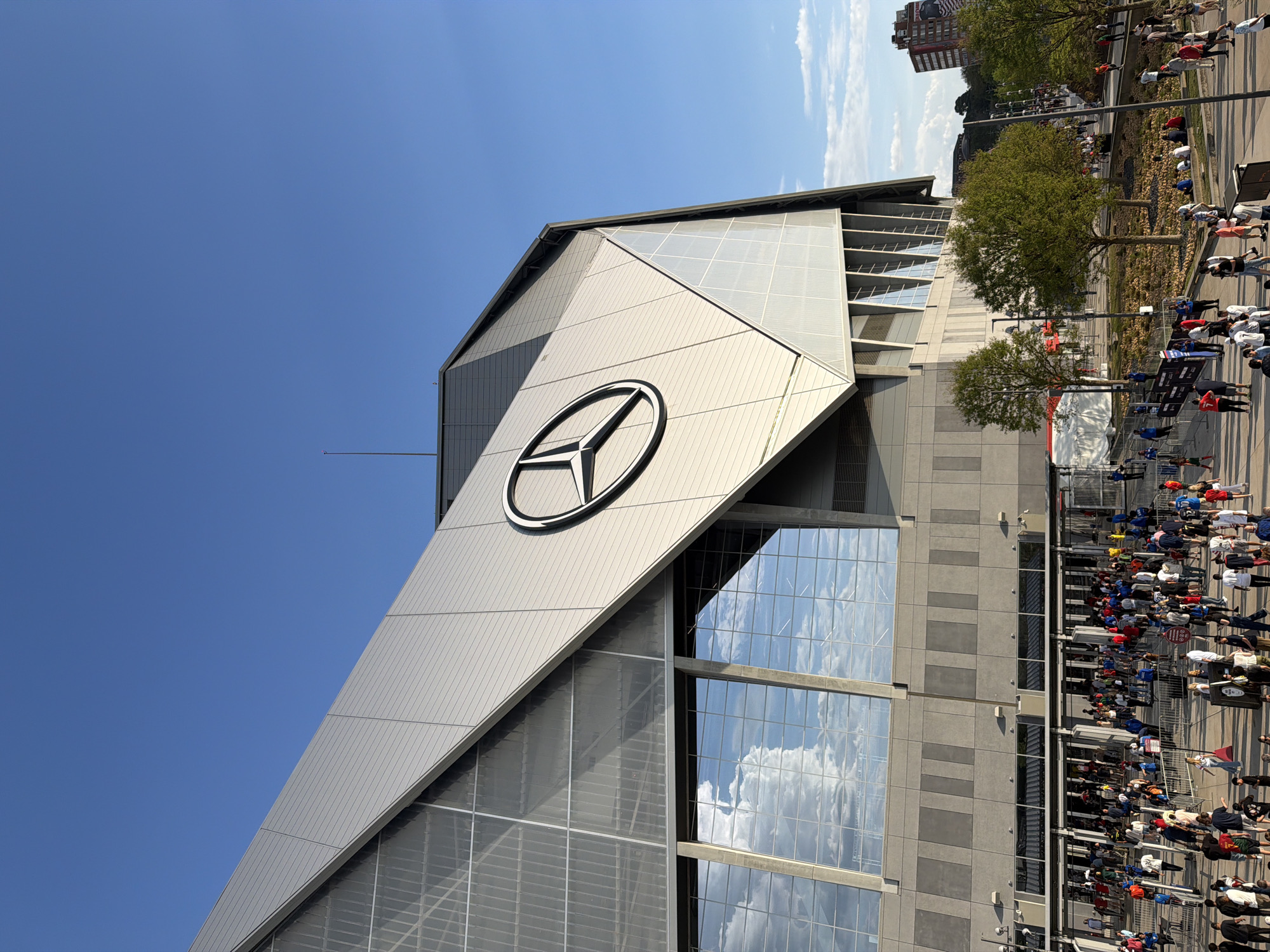The image depicts the Mercedes-Benz Stadium in Atlanta, Georgia, United States. The stadium's exterior is prominent, featuring a large Mercedes-Benz logo. The architecture includes a mix of metal panels and glass windows reflecting the sky. Crowds of people are visible in the foreground, suggesting an event or game is about to occur. Trees and a few buildings are in the background, with a clear blue sky overhead.