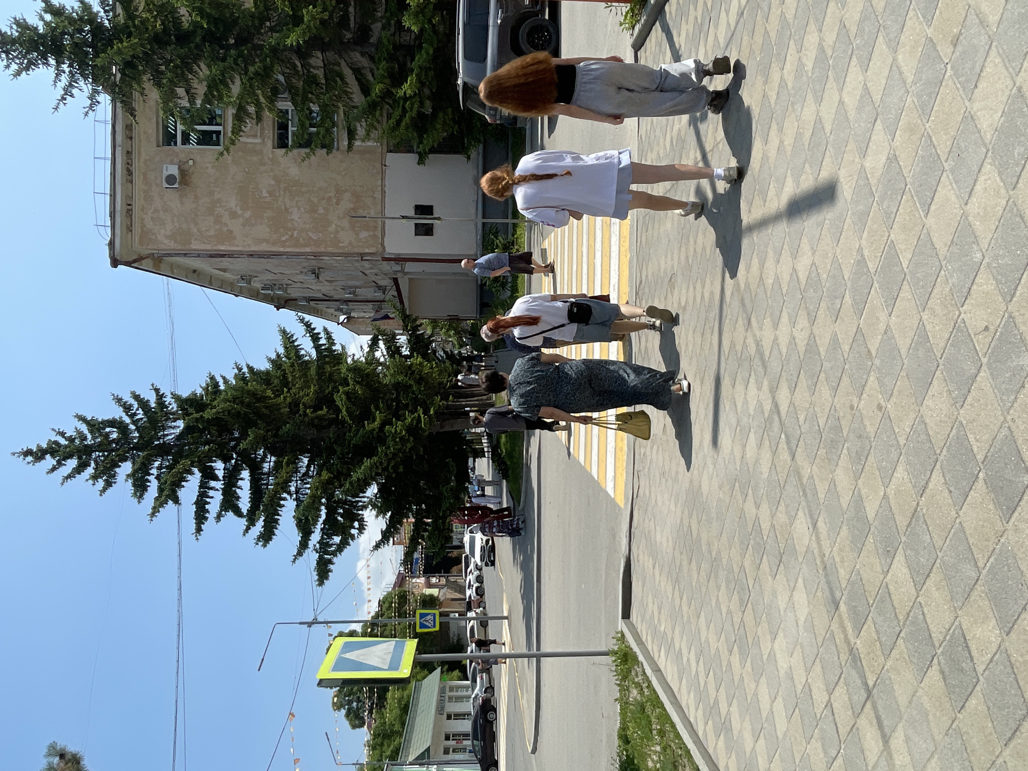 The image captures a street scene in Spassk-Dal'nij, Russia, bathed in bright sunlight, suggesting a warm, clear day. Several people are walking on a sidewalk paved with diamond-shaped tiles. In the foreground, a woman in a dark patterned dress and a yellow bag walks away from the camera, crossing a crosswalk. Behind her, a younger woman with red hair in a braid wears a white shirt and shorts. Further back, another person with long red hair walks in grey athletic pants and a black top. A man in a blue shirt and brown shorts is also visible near the crosswalk.
The setting includes a building with pale beige walls and windows, partially obscured by a large evergreen tree with dense foliage. Across the street, several cars are parked, and other buildings and trees line the road. A pedestrian crossing sign, a yellow diamond shape with a blue triangle containing a white arrow, is prominently displayed on a pole. The overall atmosphere appears to be a casual, ordinary day in a town or city. The shadows cast by the people and trees indicate it's likely midday.