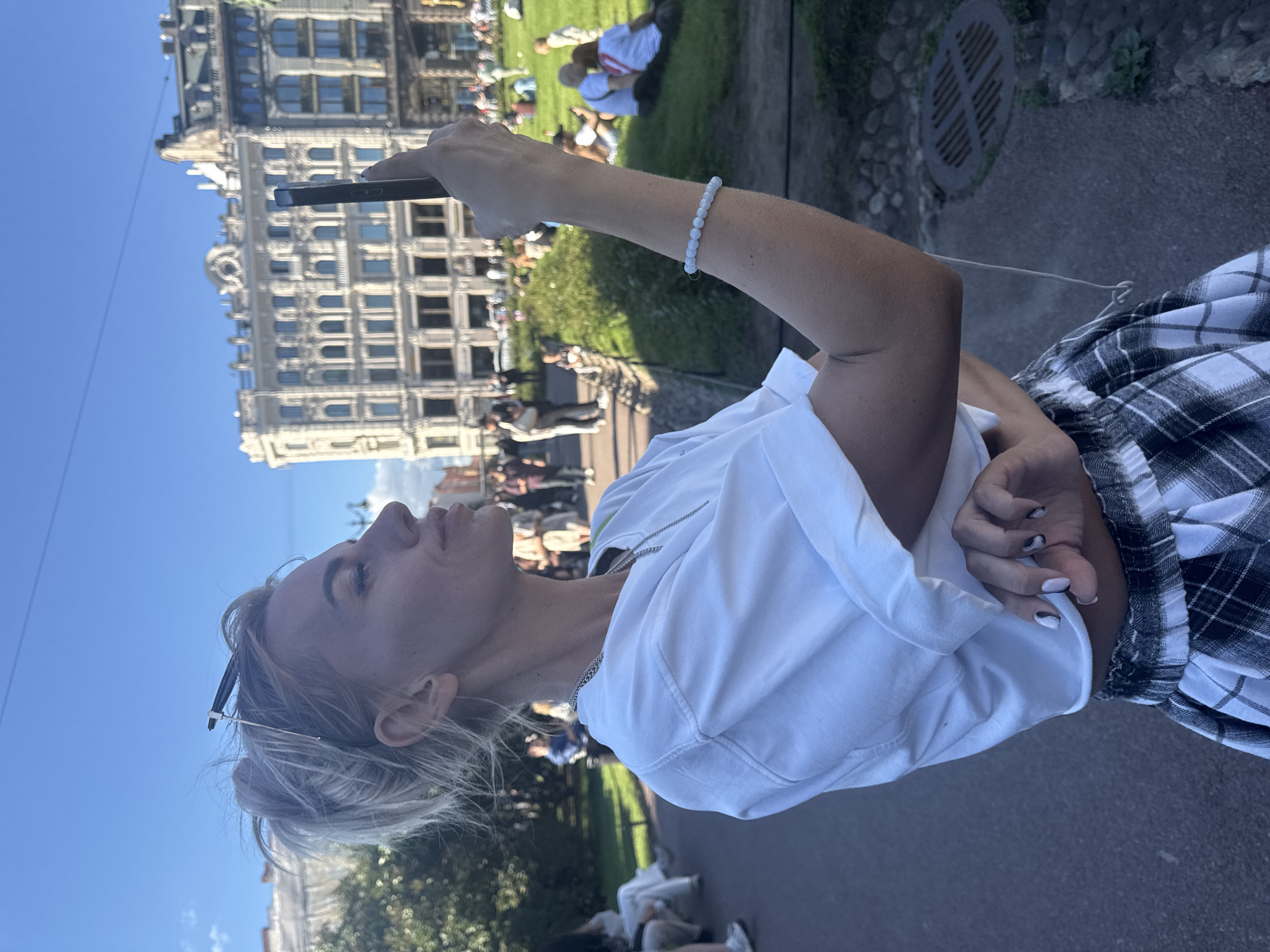 This media file depicts a young woman in Saint Petersburg, Russia, standing in front of a grand, classical-style building with arched windows. She is holding a smartphone in her right hand, seemingly taking a photo or video. The woman has blonde hair styled in a messy bun, with sunglasses perched on her head. She is wearing a white t-shirt and a black and white plaid skirt. A delicate white beaded bracelet adorns her left wrist.
The background shows a park-like area with green grass and a few other people scattered around, some sitting and others walking. The architecture of the building suggests a historic or cultural landmark within the city. The bright blue sky and ample sunlight indicate it is a daytime scene, likely late morning or afternoon, with pleasant weather for outdoor activities. The woman's pose and the way she holds her phone suggest she is engaged in capturing the moment or scenery. There is no visible text in the image.