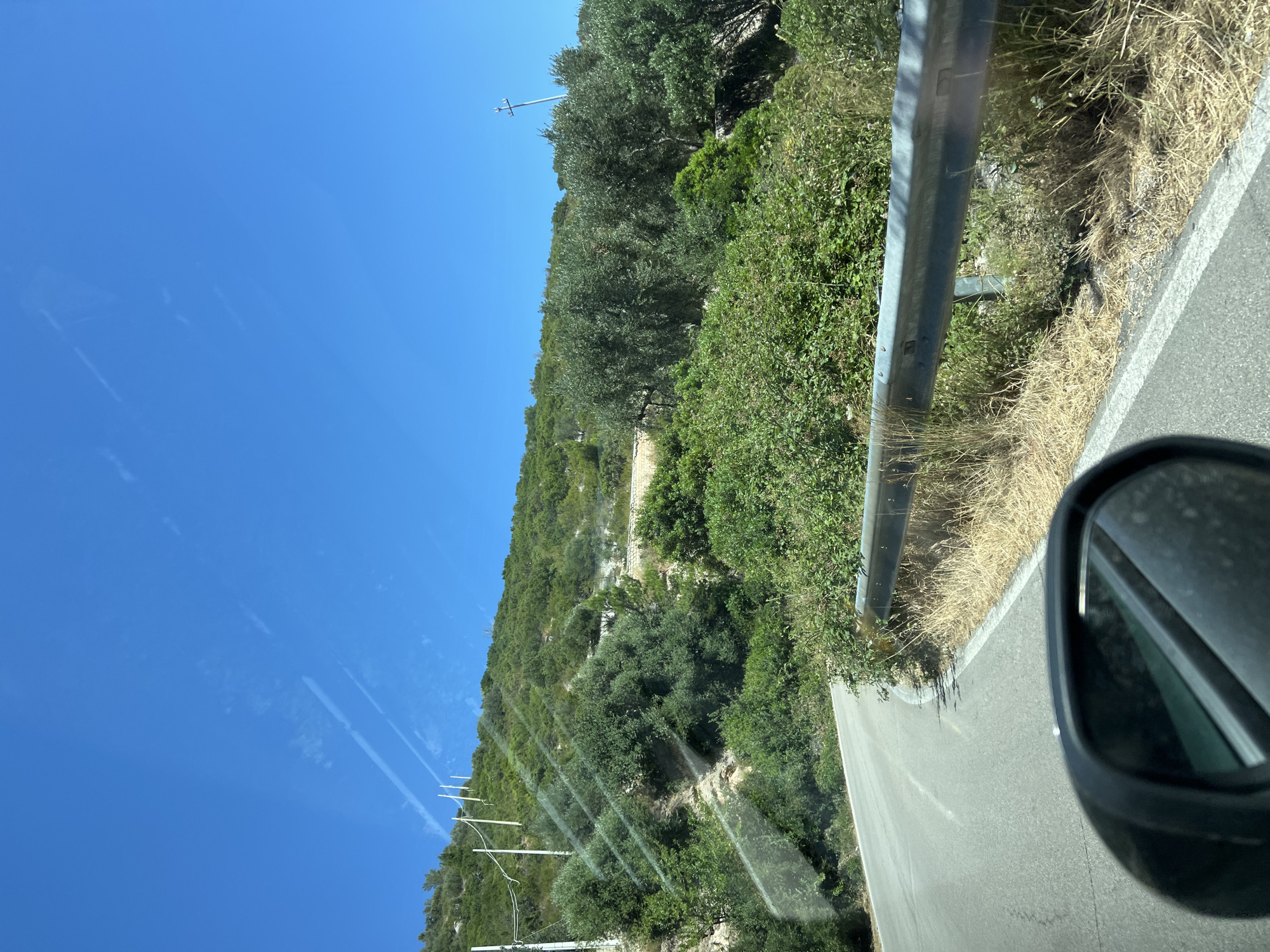 This image captures a scenic drive on a road in Mattinata, Italy, on a clear, sunny day. The road curves gently to the left, bordered by dry grass and a metal guardrail on the right. Beyond the guardrail, dense green vegetation covers the hillside. In the distance, several utility poles with wires ascend the slope, and more trees, including olive trees, are visible. The sky is a vibrant blue with a few wispy clouds. The reflection of the surrounding scenery is visible in the car's side mirror, which is in the foreground of the image.