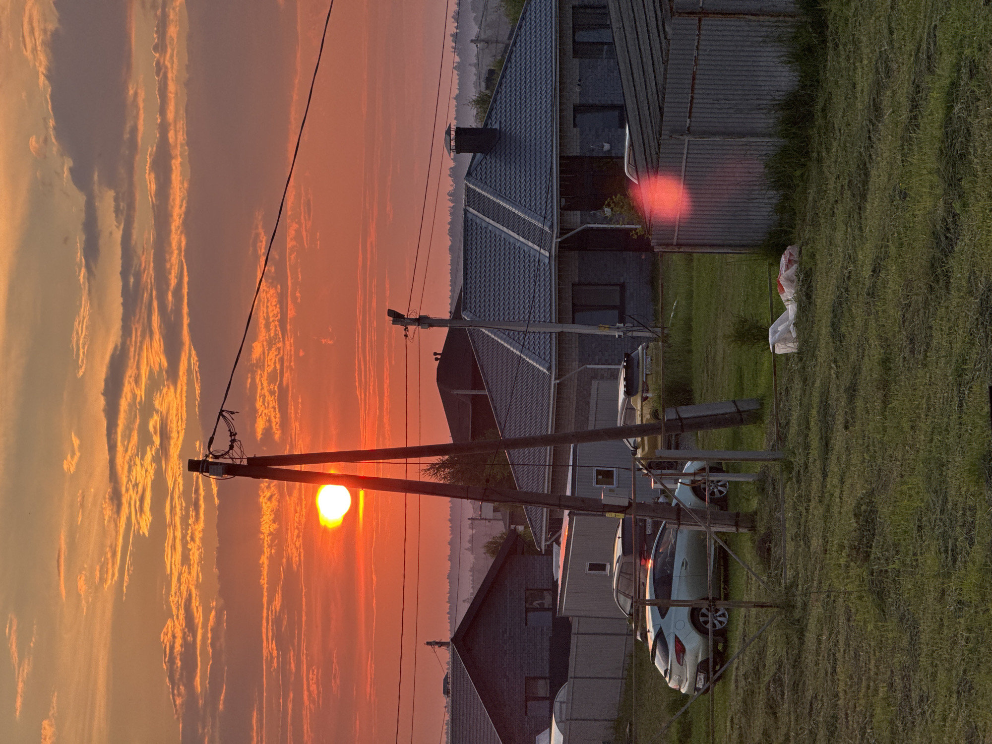 The image depicts a rural residential area at sunset. The sky is a vibrant orange and red, with scattered clouds. A bright sun is visible near the horizon, partially obscured by utility poles and wires. In the foreground, a grassy area shows signs of recent mowing. Several houses with dark-colored roofs and brick or concrete walls are visible, along with parked cars. One house has a prominent chimney. A corrugated metal fence runs across the right side of the image, and some white bags are lying on the grass near it. The scene appears to be in Izhevsk, Russia, given the presence of characteristic architecture and landscape. There are no people visible in the image.