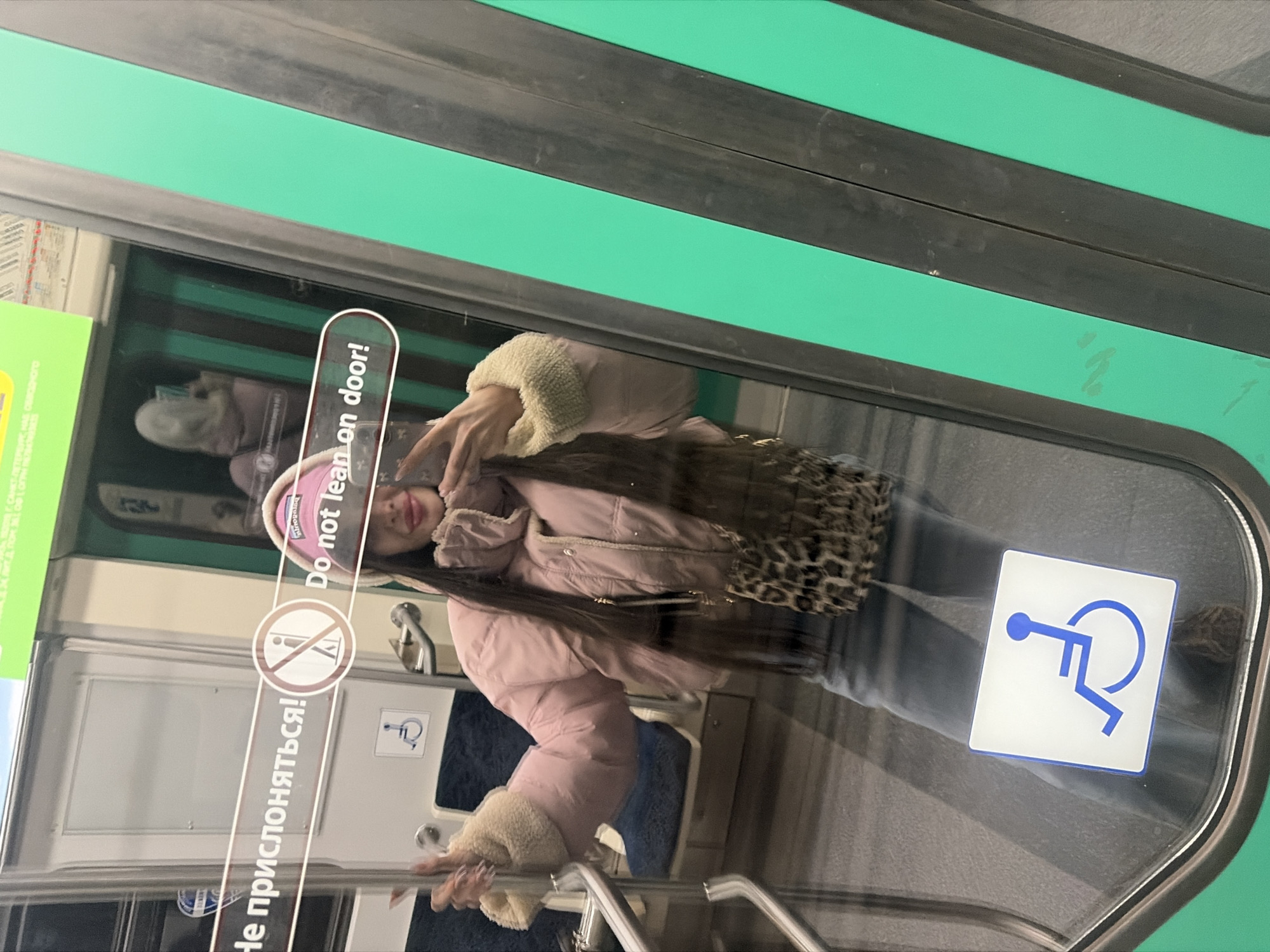 The image is taken from inside a metro train in Saint Petersburg, Russia, and is focused on the reflection of a woman in the train door. She is wearing a pink jacket, a pink hat, and a leopard print bag, and she has long brown hair. Her hand is raised towards her face. In the foreground, on the train door, are signs in Russian and English reading "Do not lean on door!" and a symbol indicating the seats are for disabled passengers. The train door is framed by metal bars. The background shows seats inside the train. The interior colors of the train are predominantly green and white.