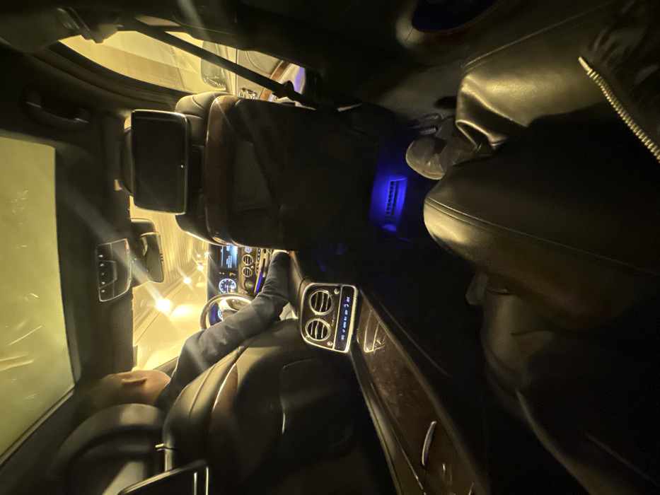 A first-person perspective photograph captured from the rear passenger seat of a luxury vehicle, confirmed as a Mercedes-Benz Maybach by the visible display, traveling through a brightly lit urban tunnel in Moscow, Russia. The vehicle's interior features dark leather upholstery, polished dark wood trim on the center console and seat backs, and subtle blue ambient lighting in the rear footwell. A male driver, visible from behind, wears a dark suit jacket and has his hands positioned on the steering wheel. The dashboard displays digital instruments and controls, while the center console features rear climate vents and a digital screen indicating "Maybach" and a temperature setting of "20 C". A black rear entertainment screen is affixed to the back of the front passenger seat. The legs of the person taking the photograph are visible in the foreground, clad in dark, possibly leather, trousers, with a light-colored shoe partially visible. The tunnel's walls and ceiling are uniformly illuminated with a warm, yellow-orange light, indicating the vehicle is in motion.