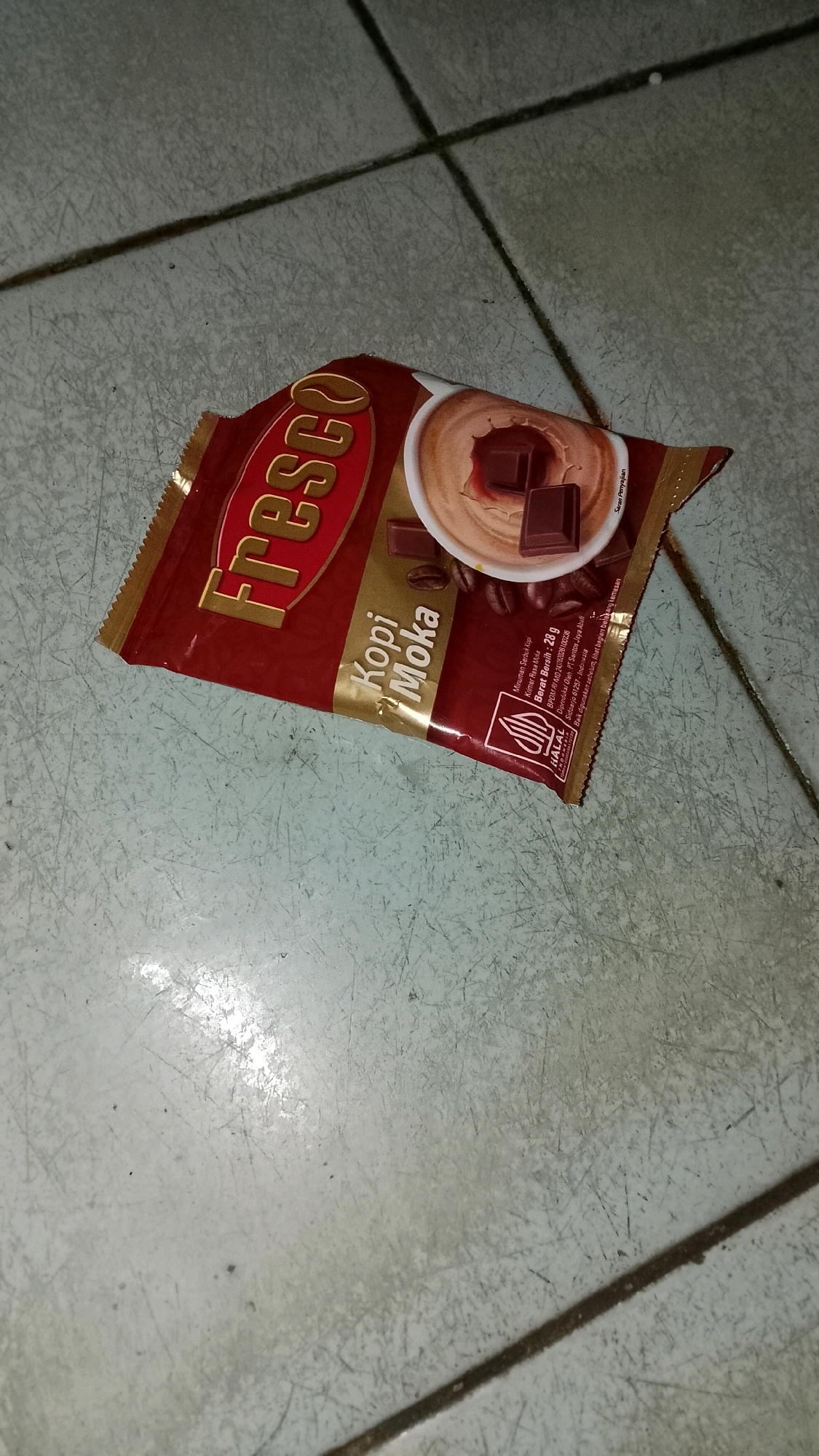 A single, unopened sachet of "Fresco Kopi Moka" instant coffee lies on a light gray, tiled floor. The sachet is predominantly maroon and gold, with the brand name "Fresco" prominently displayed in gold lettering on a maroon background. Below this, "Kopi Moka" is written in white text with a brown shadow. The image on the sachet depicts a cup of frothy coffee with chocolate pieces. Nutritional information and a HALAL certification logo are visible on the lower right side of the sachet. The tiled floor has visible grout lines and appears somewhat scuffed. The lighting is dim, casting shadows and highlighting the texture of the floor. The scene appears to be indoors, with no indication of the specific location within Mulyo Haji, Indonesia.