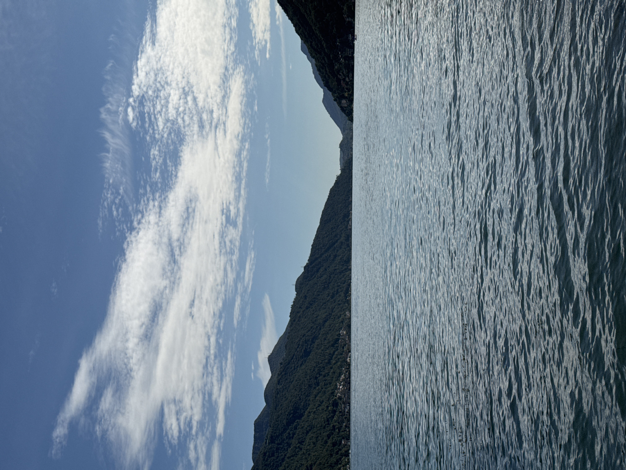 This image captures a scenic view of Lake Lugano in Melide, Switzerland. The foreground is dominated by the rippling surface of the deep blue water, reflecting the sky and surrounding landscape. The middle ground features the lush, green slopes of mountains that rise dramatically from the lakeshore. Small clusters of buildings, indicative of a town or village, are visible nestled on the mountainside. In the distance, the lake narrows, with more mountains receding into the haze under a clear blue sky dotted with wispy white clouds. The overall impression is one of natural beauty and tranquility.