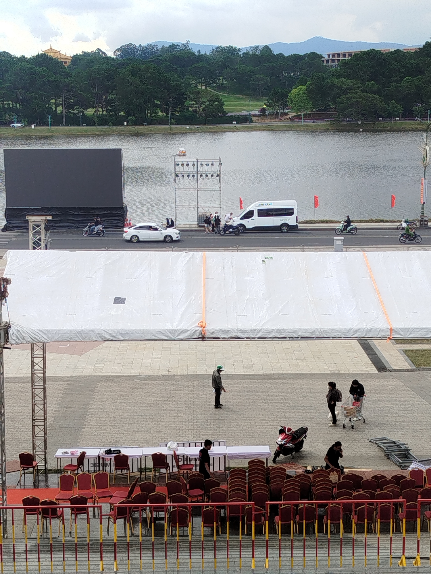 The image captures a scene near Xuan Huong Lake in Da Lat, Vietnam. In the foreground, a set of red chairs with gold trim are arranged on a tiered platform, some of which are stacked. Beyond the chairs, there are white tables set up with a few people walking around. In the middle ground, a large white tent or covering is erected, with orange straps visible. Further back, a body of water, likely Xuan Huong Lake, stretches across the frame, with a large black screen on the left and a road with vehicles and people on the right. The background features lush green trees and buildings in the distance, with a cloudy sky overhead. Several individuals are seen walking or operating vehicles, and a shopping cart is present near some of the people in the middle ground. The overall impression is one of an outdoor event or preparation for an event.