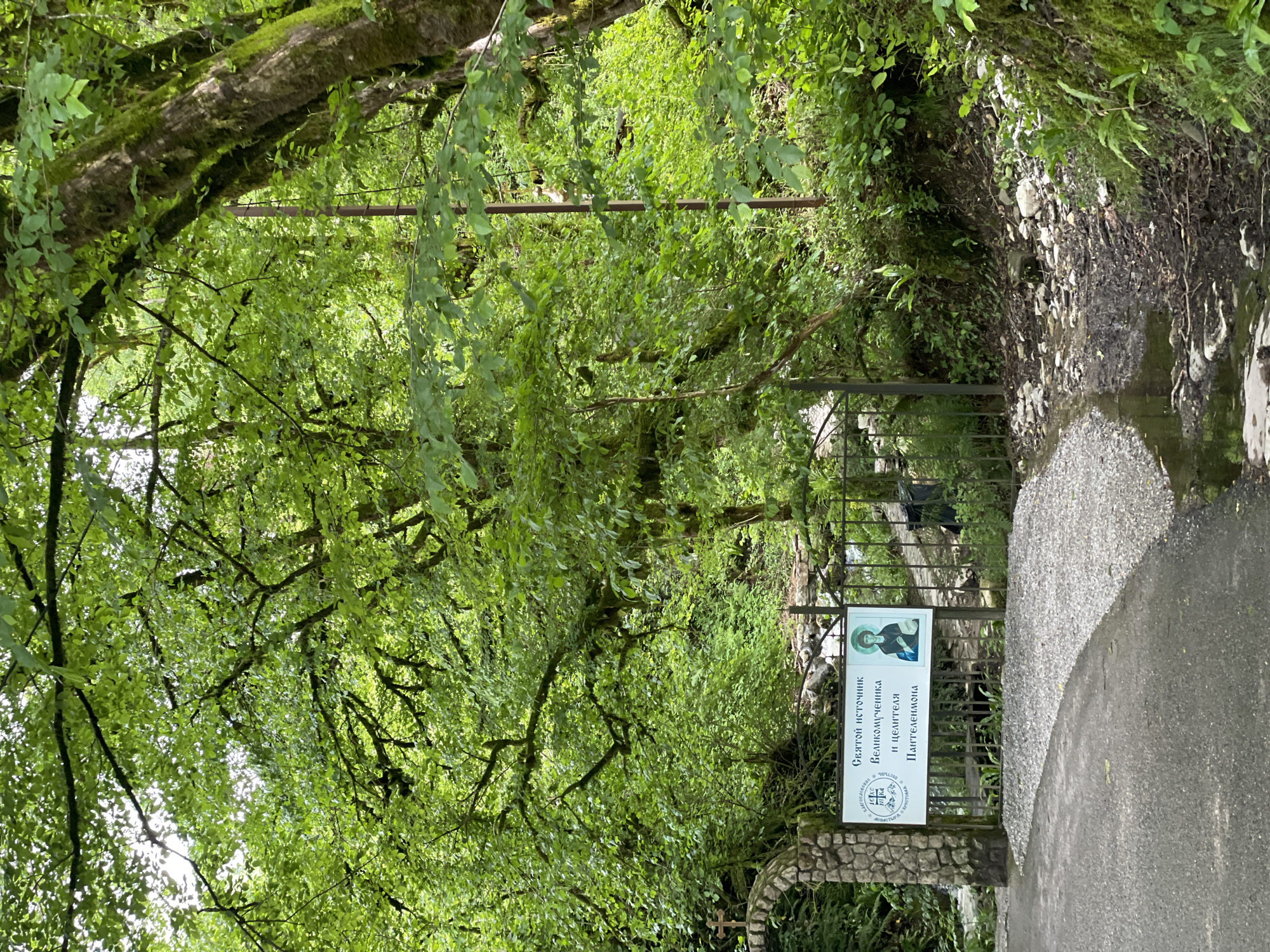 The image shows a path leading to a religious site in Solokh-Aul, Russia. The path, partially paved, is flanked by dense green foliage. A sign, inscribed in Russian and featuring an image of a religious figure, stands at the entrance. A low stone archway is visible. The scene is enveloped by trees, creating a shaded environment.