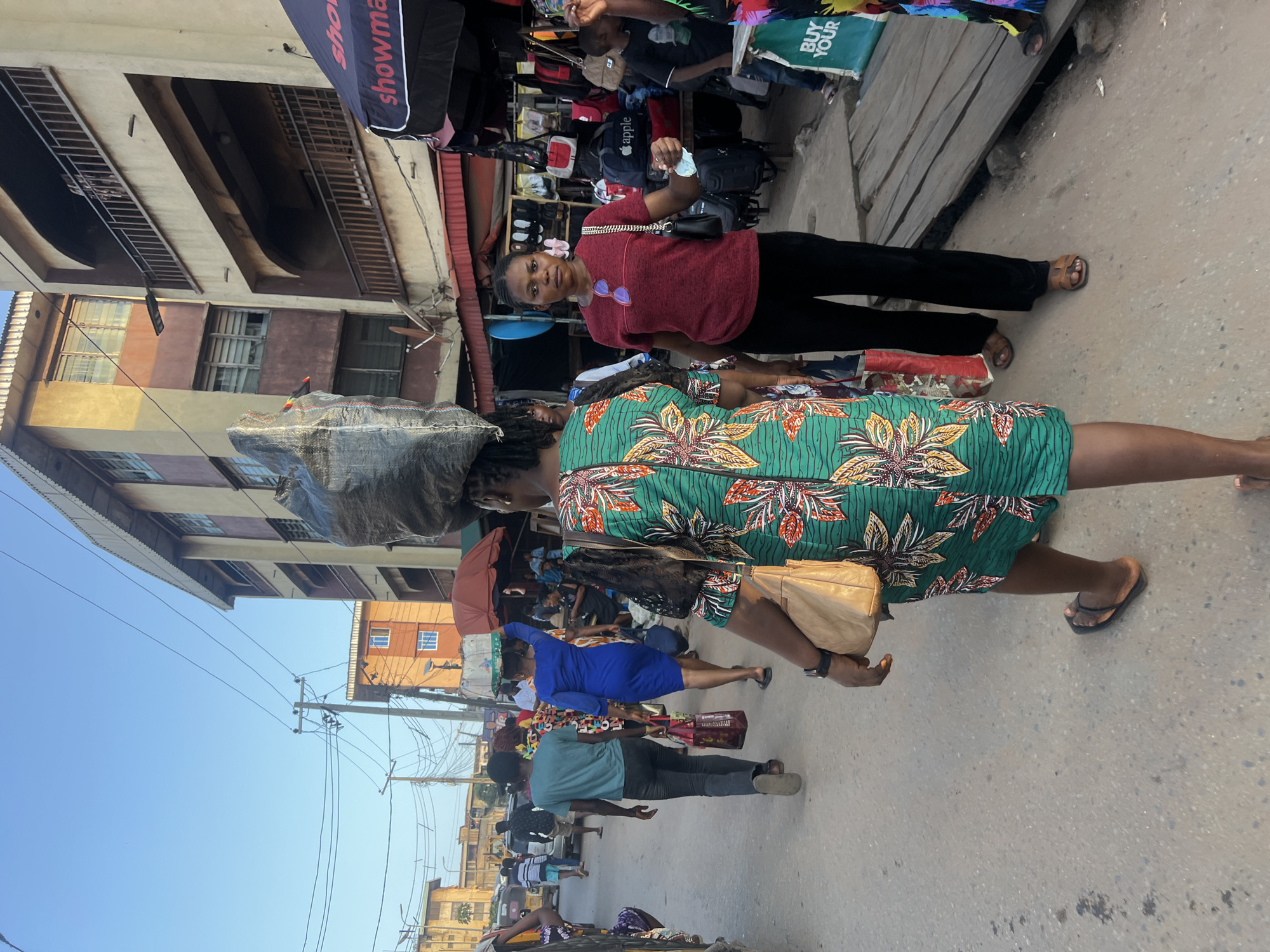A medium shot captures an outdoor street scene in Nkpor, Nigeria, during the day. In the foreground, two women walk away from the camera. The woman on the left wears a vibrant green dress with a floral pattern and carries a tan shoulder bag. The woman on the right is partially obscured by the woman on the left, wearing a black top and black pants, with her left hand held out. Behind them, a crowded street unfolds with various individuals walking, and on the right side, market stalls are visible. Buildings line both sides of the street, some appearing to be multi-story structures with balconies. Power lines and poles are present, indicating an urban setting. The ground is paved and appears somewhat worn. The sky is clear and blue.