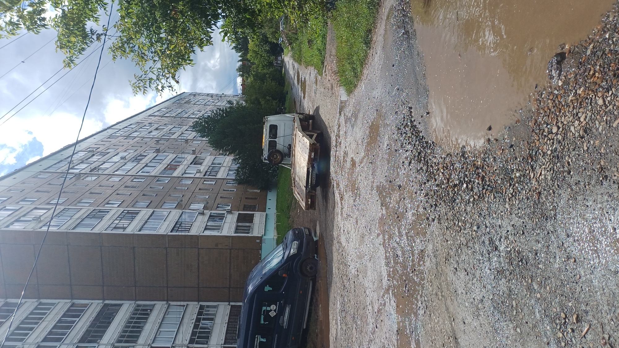 The image shows a muddy, gravelly road in front of a multi-story apartment building in Izhevsk, Russia. A dark-colored minivan is partially submerged in a large puddle on the left side of the frame. To the right of the minivan, a white tow truck with a flatbed is parked. The building appears to be constructed of concrete panels and has many windows. Trees and other vegetation are visible along the side of the road. The sky is cloudy. The scene depicts a post-rain or poorly drained urban environment.