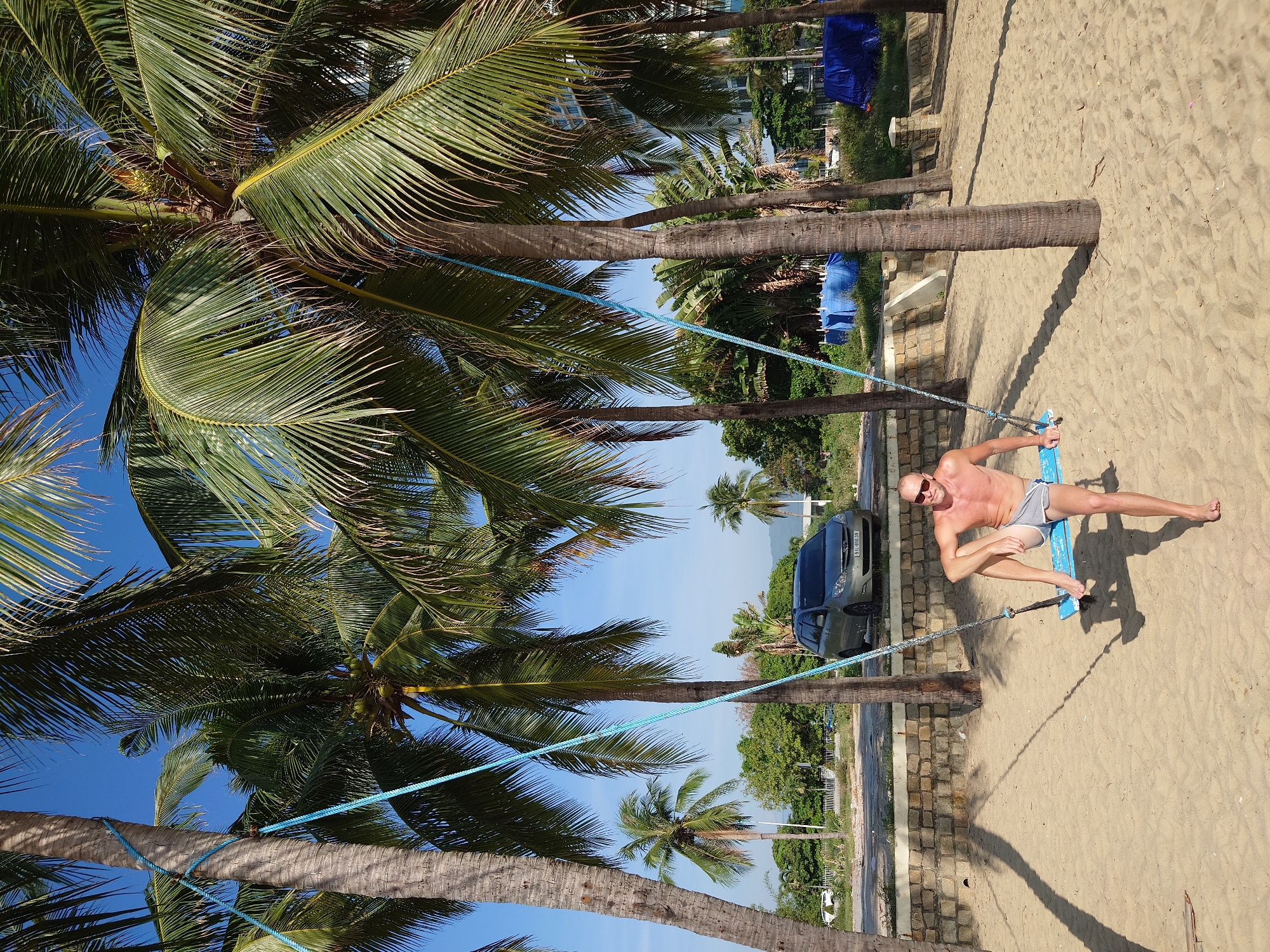 A shirtless man with sunglasses is sitting on a blue swing under palm trees on a sandy beach. He is wearing grey shorts. The swing is suspended by blue ropes tied to the palm trees. A silver minivan is parked behind a low stone wall in the background, along with some greenery and buildings. The sky is clear and blue.