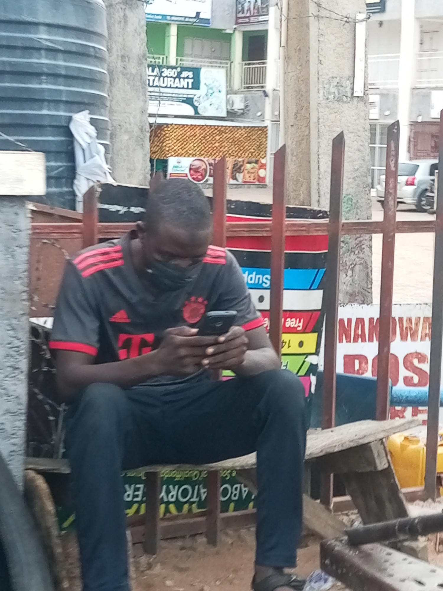 The image shows a man sitting outdoors, engrossed in his smartphone. He is positioned on a simple wooden bench or structure, with a large black water tank on his left and metal poles forming a barrier in the foreground and midground. The man wears a dark grey short-sleeved t-shirt with red stripes on the shoulders, an Adidas logo, and a "T" logo (resembling the FC Bayern Munich kit). He is also wearing dark trousers, sandals, and a dark face mask covering his lower face. His head is bent, and both hands hold his phone, indicating he is focused on its screen.
The setting appears to be an urban or semi-urban street in Tempure, Nigeria, with multi-story buildings in the background housing various businesses. Visible text on signs includes "LA 360° JPS TAURANT" (Restaurant), as well as partial words like "AKOWA," "DOS," "Free," "14," and "Supp." Inverted text such as "ABO ATORY ER" (likely "LABORATORY") and "e Soof Qualiffarda Orimer" is visible on a green sign directly below where the man is seated. A white car is parked in the distance on the right, and yellow jerrycans are seen in the lower right corner. The lighting suggests it is daytime, possibly overcast or during the early morning/late afternoon hours.