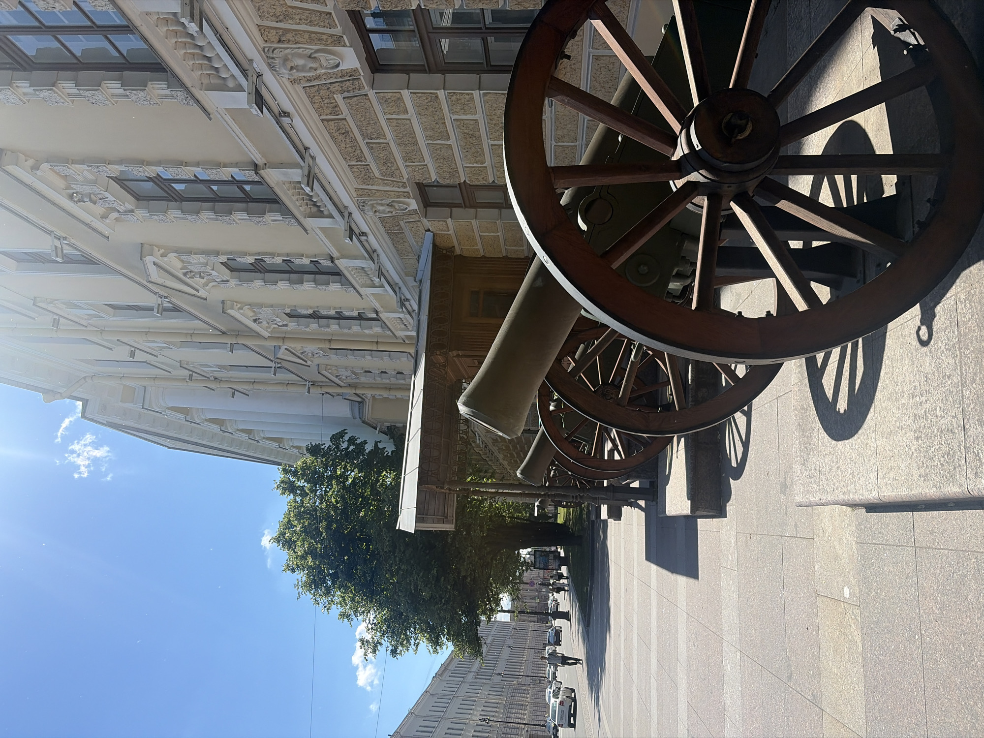 The image depicts a street scene in Saint Petersburg, Russia, likely in front of a museum or historical building. In the foreground, several old cannons are displayed on a sidewalk, their wooden wheels and metal barrels casting shadows on the paved ground. The cannons are aligned facing away from the viewer, suggesting they are part of an outdoor exhibition. Behind the cannons, a grand, classical building with ornate architectural details, including columns and decorative window frames, rises up. A large green tree with dense foliage stands to the left of the cannons, providing a natural contrast to the man-made structures. Further down the street, a row of similar classical buildings lines the left side of the road, with cars and a few pedestrians visible. The sky is a clear blue, with a few wispy clouds and the sun shining brightly, indicating a sunny day. The overall impression is one of a historical and cultural urban setting.