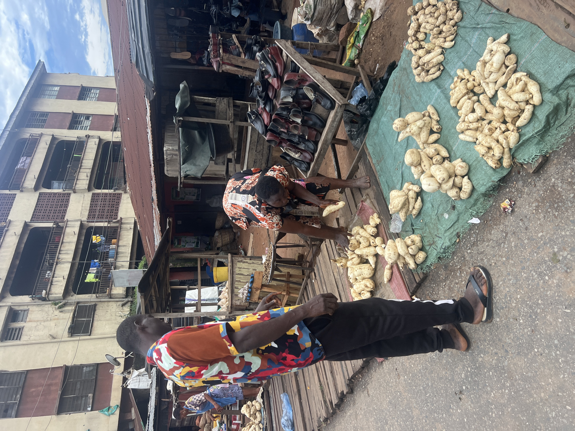 This is a photograph taken at the Nkpor Main Market in Nkpor, Nigeria, on a cloudy day. In the foreground, a man wearing a colorful, abstract-patterned shirt and black pants with white stripes stands on a wooden plank walkway. He is facing left, looking down. To his right, on a green tarp spread over wooden planks, is a large pile of yams. Another person, wearing a dark patterned dress, is seen from the waist down, bent over tending to yams on the tarp, with one yam in their hand. In the background, stalls with corrugated metal roofs are visible. One stall displays a variety of shoes. Further back, a multi-story building with balconies and clothes hanging from them overlooks the market. The overall scene depicts a bustling market environment.