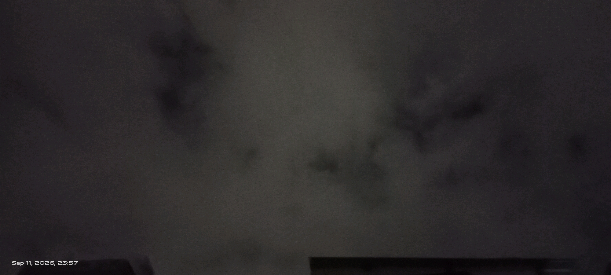 The image is a dark, grainy photograph taken at night, indicated by the timestamp "Sep 11, 2026, 23:57". The scene appears to be outdoors, with a predominantly dark and cloudy sky. There are indistinct shapes visible, likely clouds or some form of atmospheric haze, with darker patches scattered throughout. In the lower portion of the image, a dark horizontal structure is partially visible, suggesting the presence of a building or some man-made object. The lower-left corner shows the text "Sep 11, 2026, 23:57", which is likely a timestamp embedded by the camera. There are no people, animals, or clear objects identifiable in the image, beyond the very faint hints of structures. The image quality is poor due to low light and potential motion blur. The location of Daria, India is mentioned, but there are no visual elements in the image that can confirm this specific location.