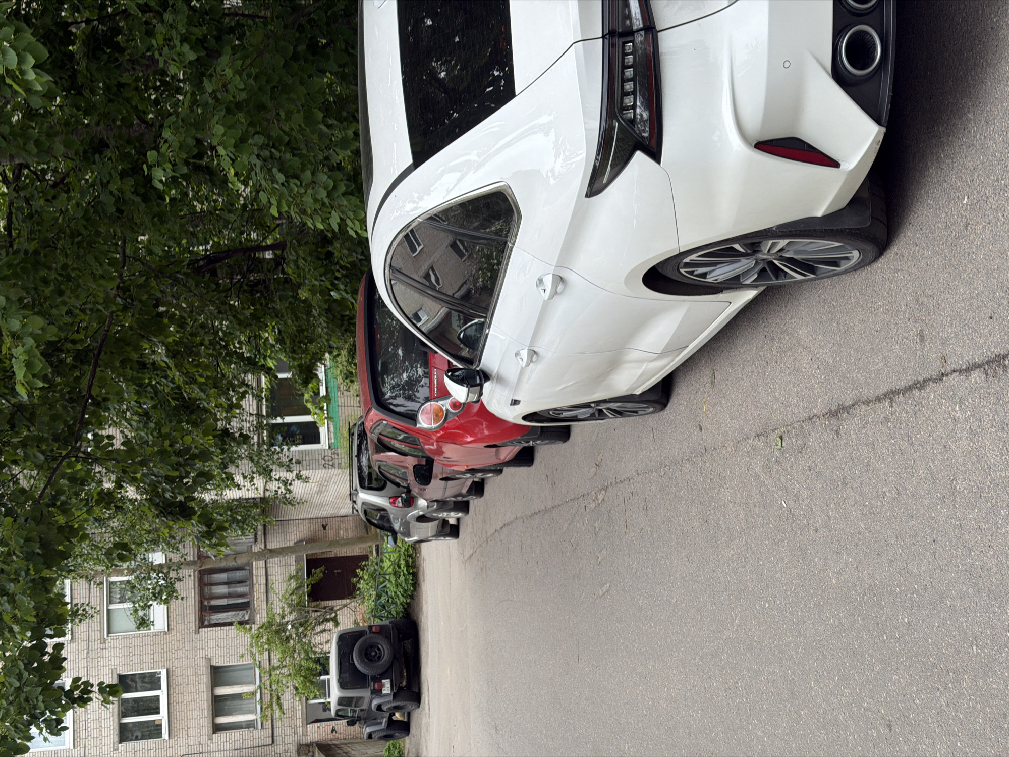 The image captures a static street scene in what appears to be a residential area of Novosergievka, Russia, during bright daytime conditions, possibly under an overcast sky.
A line of vehicles is parked on an asphalt surface, extending into the distance. In the foreground, a white modern sedan or crossover is prominently featured, showcasing its rear and side profiles. This car has a distinctive design with black-tinted rear lights, dual chrome exhaust tips, and multi-spoke alloy wheels. Behind it, a red hatchback is parked, followed by a gray hatchback or crossover, and then another dark sedan. At the far end of the row, closest to the building, a dark-colored SUV, resembling an older off-road model with a spare tire mounted on its rear, is visible.
To the left of the parked cars, dense green foliage from large trees covers a significant portion of the scene, partially obscuring a light-colored brick residential building. The multi-story building features several windows, some with modern white frames and others with older, darker frames. A dark entrance door is visible on the ground floor. Faint, weathered text or numbers, possibly "202," are discernable on the building's facade near the ground level, partially hidden by tree branches. The street is paved with asphalt, showing some minor wear. No people or active events are depicted; the scene is one of quiet, parked vehicles in an urban setting.