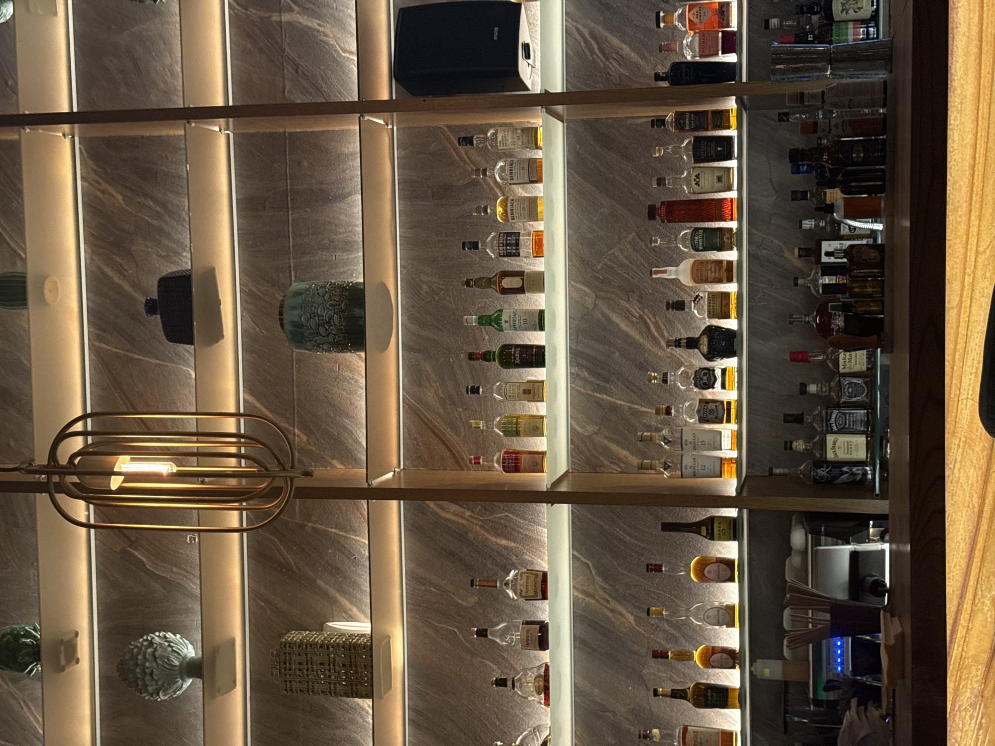 The image displays a bar with multiple shelves stocked with bottles of liquor, predominantly whiskey, cognac, and other spirits. The bottles are arranged in rows, with several brands and varieties visible. The bar shelves are illuminated with vertical lights. Additional decorative items are on the shelves, including vases and a modern-style light fixture. The scene appears to be in a well-lit indoor environment, potentially a bar or restaurant. No people are visible. The location and city are unknown based solely on the image.