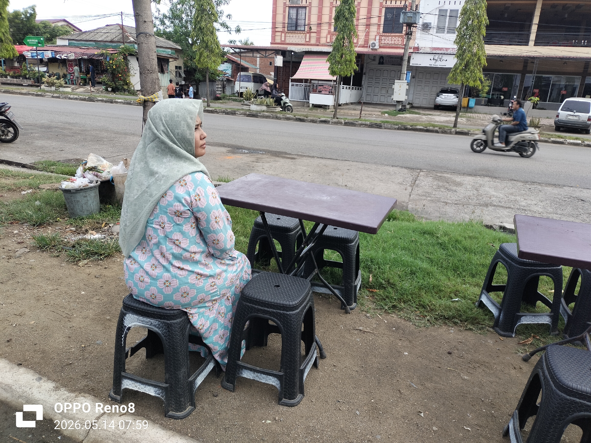 The photograph depicts a woman seated on a plastic stool at a roadside establishment in Lhokseumawe, Indonesia. She is wearing a light green hijab and a light blue floral patterned outfit. She is positioned next to a dark brown plastic table supported by three plastic stools, one of which she occupies. Another identical table and stool set is visible nearby. The ground is partially covered with dirt and grass. A street is visible in the background with a motorbike, cars, and buildings lining the sides. The time stamp on the image indicates May 14, 2026, at 07:59.