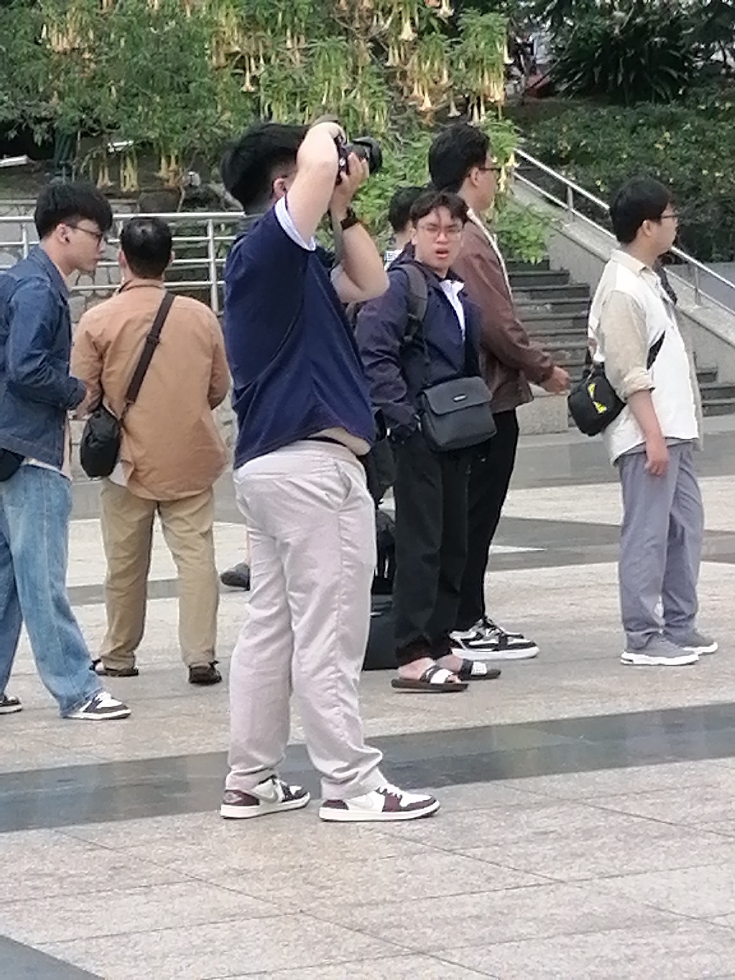 The image shows a group of five Asian men in an outdoor urban setting, likely a public plaza or walkway. One man in the foreground, wearing a dark blue shirt and light gray pants, is facing away from the camera and appears to be taking a photograph with a black camera. He is wearing white and black sneakers. To his left, another man in a light brown shirt and khaki pants is also facing away, with a dark bag slung over his shoulder. He is wearing sandals. To the left of these two, a man in a denim jacket and blue jeans is partially visible, also facing away and wearing sneakers.
Further into the background and to the right, a man in a dark jacket and black pants is standing and looking to his right. He is wearing black and white sneakers and carrying a dark gray shoulder bag. To his right, another man in a light-colored shirt and gray pants is standing and looking to his left. He is wearing gray sneakers and a black bag is visible on his shoulder.
The environment includes a paved ground surface, a set of stairs in the background with a metal railing, and abundant green foliage with yellow flowers visible in the upper portion of the image. The lighting suggests it is daytime. There are no specific visual cues that definitively place the location in Da Lat, Vietnam, such as distinct architecture, signage, or recognizable landmarks.
