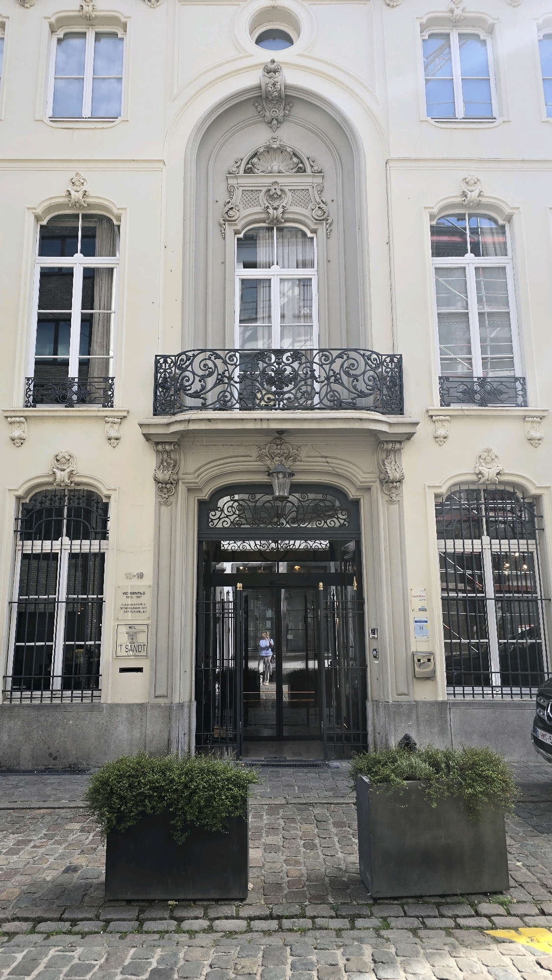 The image shows the exterior of a building in Antwerp, Belgium. The building has a light-colored facade with ornate architectural details, including a balcony with decorative ironwork above the entrance. The entrance itself is a large, glass-enclosed doorway with metal gates. In front of the entrance are two large rectangular planters filled with green foliage. The ground in front of the building is paved with cobblestones. A person is visible through the glass doors, appearing to be exiting the building while riding a bicycle. There is a sign on the building that reads "HOTEL T SANDT" and another sign with the numbers "13-19". The overall scene depicts a building entrance on a street, with elements of both historical architecture and modern design.
