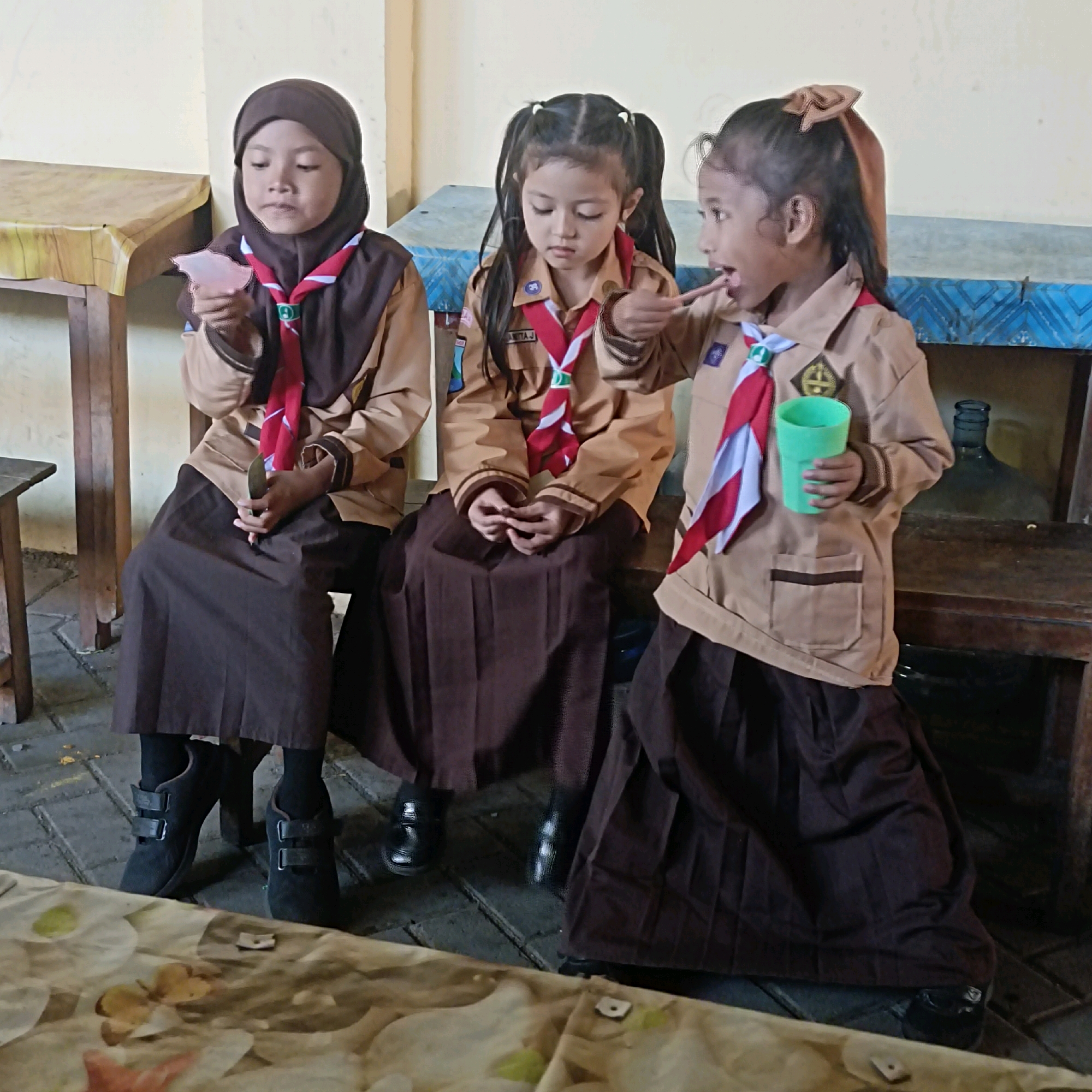 Three young girls in scout uniforms are sitting on a bench in what appears to be an outdoor or semi-outdoor setting. The girl on the left is wearing a brown hijab and holds a small pink object in her hand. The middle girl has her hair in pigtails and is looking down at her hands, which are clasped in her lap. The girl on the right is facing left and appears to be talking or interacting with the middle girl, holding a green cup in her right hand and a thin object, possibly a stick or toothpick, which she is using to gesture towards the middle girl's mouth. All three girls are wearing brown skirts and brown scout jackets with red and white neckerchiefs. The background shows wooden furniture and a large glass bottle, suggestive of a rustic or educational environment. The floor is paved with bricks. The overall scene depicts a moment of interaction among the scouts.
