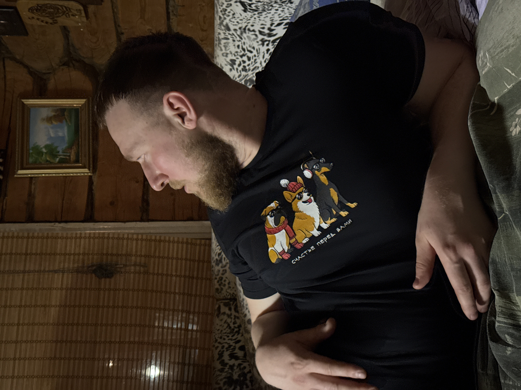 A man lies on a bed in a room with wood paneling on the walls. He is wearing a black t-shirt featuring a graphic of three dogs and Russian text. The room contains a small framed picture on the wall and a patterned bedspread. The location is potentially Rosstani, Russia.