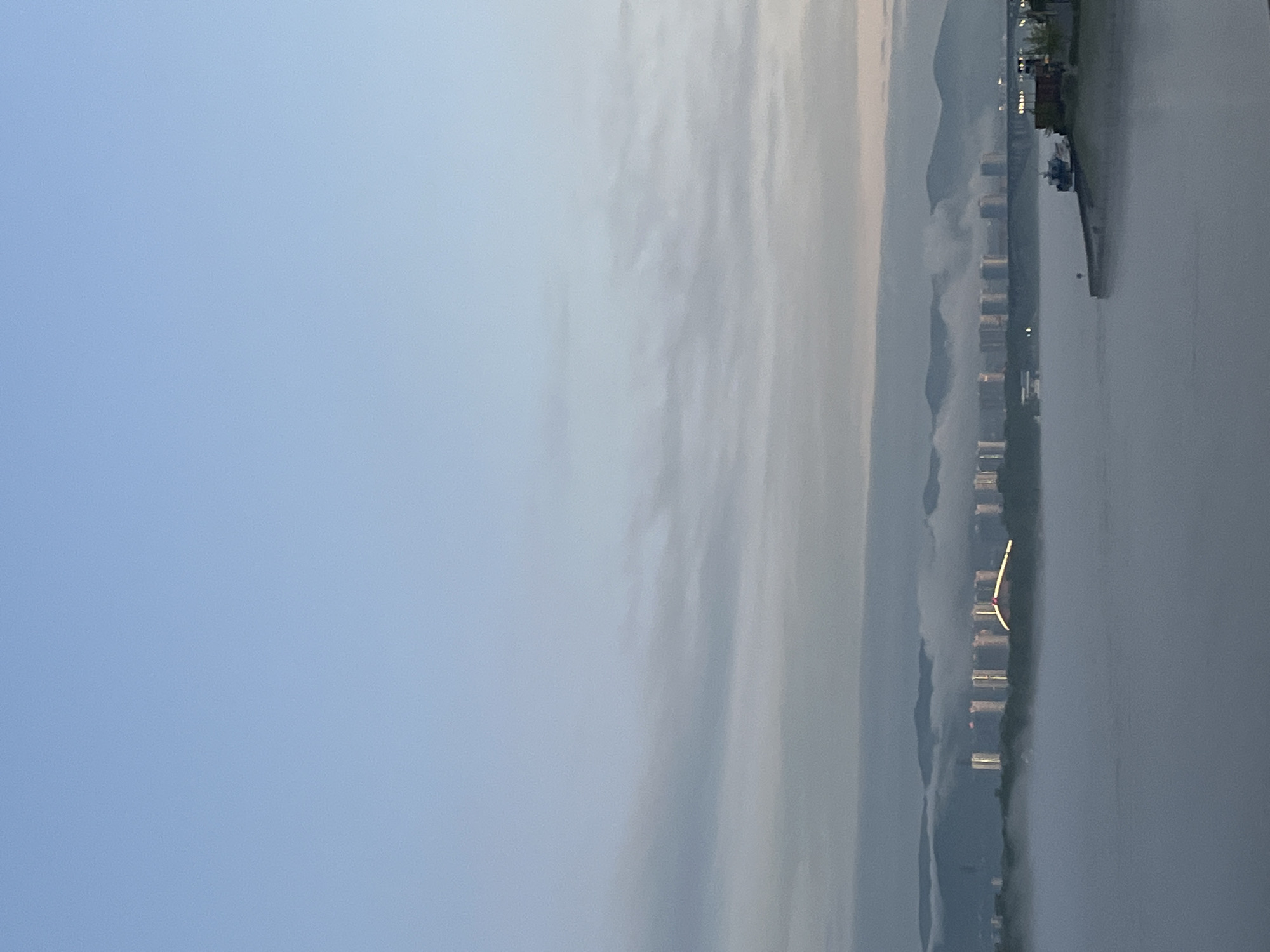 The image captures a cityscape along a body of water, possibly the Yenisey River in Krasnoyarsk, Russia, given the presence of hills and characteristic high-rise buildings along the waterfront. The scene has a misty or overcast sky, with light reflecting on the water. There is a pier visible along with some boats. The picture appears to have been taken at dawn or dusk based on the soft lighting.