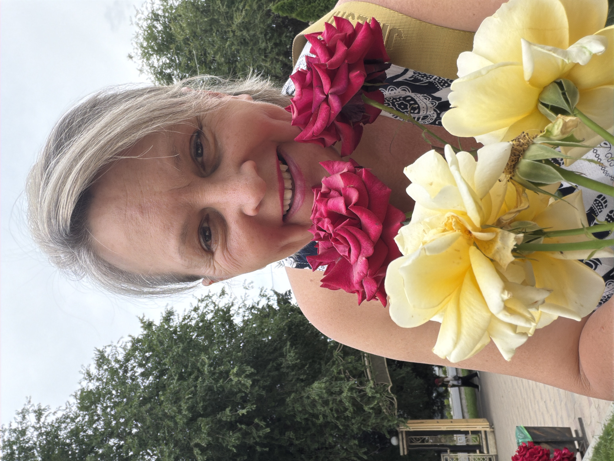 A medium close-up shot features a woman with fair skin and short, graying blonde hair, looking directly at the camera with a wide smile. She is holding a bouquet of roses, with two dark red roses in the upper part of the frame and several pale yellow roses in the foreground. The woman is wearing a dark and white patterned dress and a mustard-yellow strap over her shoulder. The background is slightly blurred, showing green trees and a grey, overcast sky. To the left, a decorative archway with golden accents and a trash bin are visible. Further in the background, a person is walking on a paved path.