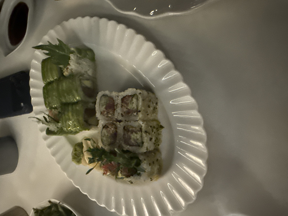 A white, oval ceramic platter with a scalloped rim occupies the central frame, resting on a light-colored table in an indoor dining setting within Dubai, United Arab Emirates. The platter holds twelve pieces of sushi. Six pieces are maki rolls, wrapped in thin green vegetable strips, likely avocado, with a visible dark red filling, consistent with tuna, and a lighter creamy substance. These are garnished with small green leaves. The other six pieces are rice-outside rolls, speckled with dark sesame seeds or nori flakes, also containing a dark red filling and a lighter creamy component, topped with a few small red garnishes and green herbs. On the platter, alongside the sushi, are a compact mound of green paste (wasabi) and several thin, pale yellow slices (pickled ginger). Partially visible objects surrounding the main platter include a segment of a small white bowl containing green food items, a dark-colored smartphone, and a small white dish holding a dark liquid (soy sauce). No individuals are present, and no actions or interactions are depicted. The environment is characterized by soft, indirect lighting.