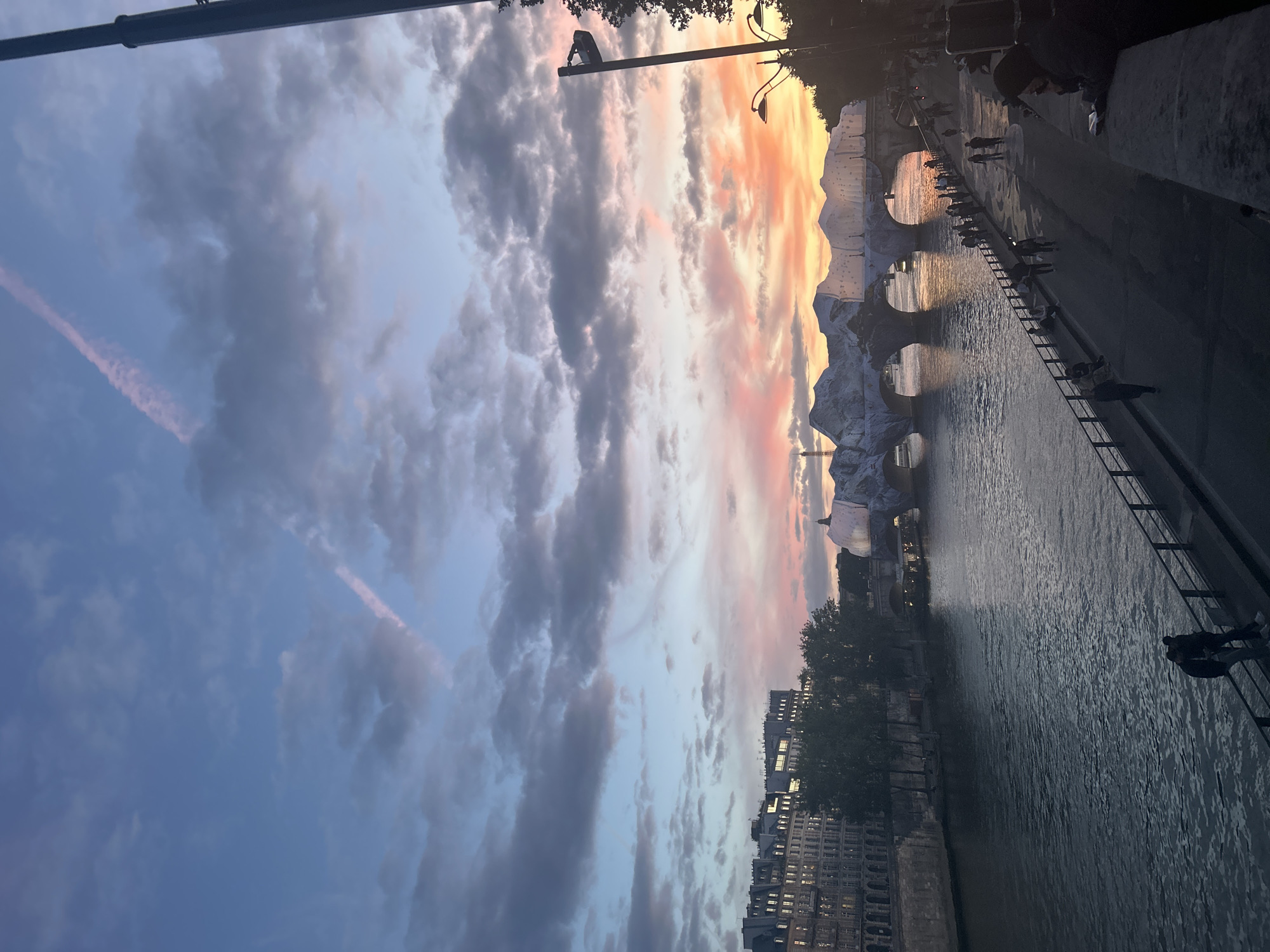 The image depicts a scenic view of the River Seine in Paris, France, during sunset. The sky is painted with hues of orange, pink, and blue, with scattered clouds. In the background, a bridge spans the river, adorned with a unique temporary structure that resembles snow-capped mountains or ice formations, possibly part of an art installation or event. The Eiffel Tower is faintly visible in the distance to the left. Along the riverbank, a paved walkway is lined with people enjoying the view. Streetlights and trees are also visible along the promenade. The water of the Seine reflects the colors of the sky, adding to the tranquil atmosphere. The architecture of the buildings on the left bank of the river suggests a historical Parisian setting.