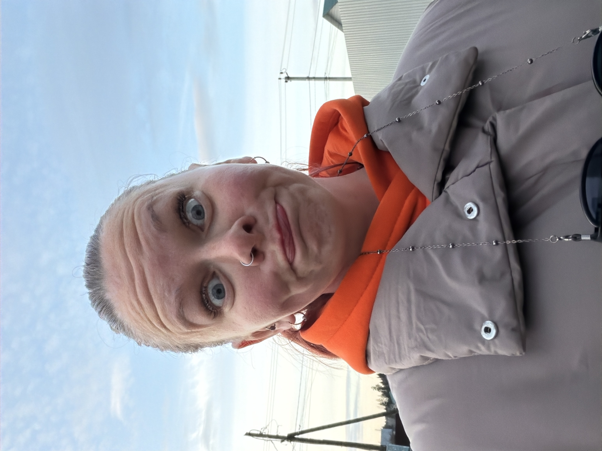 A selfie of a woman with light skin, blue eyes, and grey hair pulled back, taken at an angle looking up towards a bright blue sky with wispy clouds, possibly in Chaikovsky, Russia. She is wearing an orange hoodie under a grey puffer jacket. She has a silver nose ring and multiple earrings. A metal chain with small beads is draped across her jacket. Behind her are utility poles and buildings.