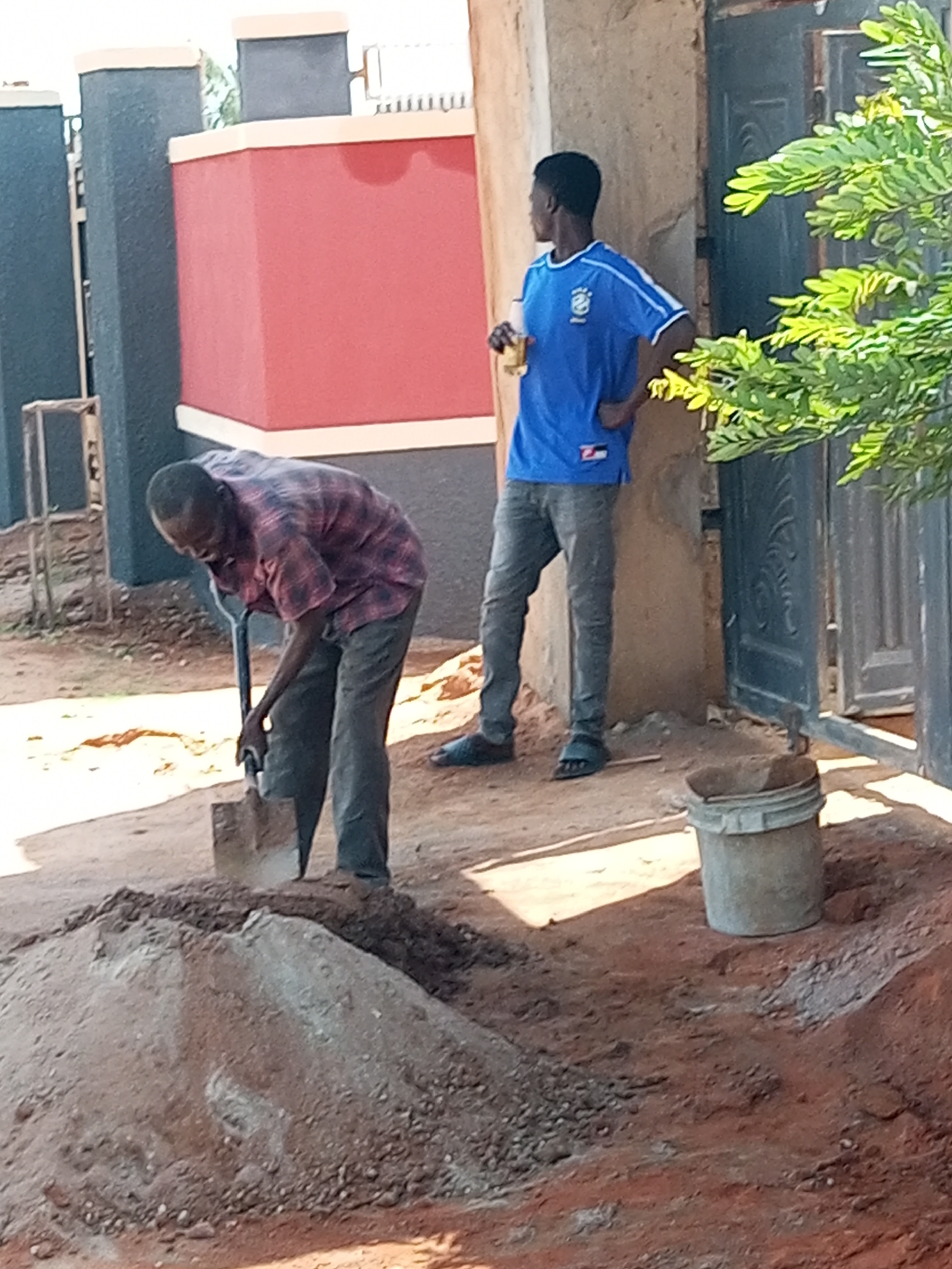 Two men are engaged in construction work in what appears to be a residential area in Tempure, Nigeria. One man, wearing a red and black plaid shirt and dark pants, is actively shoveling a pile of dirt. The other man, younger, is dressed in a blue t-shirt with white stripes and grey pants, standing with a drink in his hand. He has his hand on his hip, observing.
The scene is outdoors, with a building under construction or renovation. A red wall with white trim is visible in the background, as are dark pillars and a dark metal gate. Green foliage from a tree overhangs the scene on the right side. The ground is covered in dirt and construction debris, with a large mound of soil being worked on. A grey bucket sits to the right of the dirt pile. The lighting suggests it is daytime, likely sunny given the shadows. The overall mood appears to be one of work and casual observation. There is no visible text in the image.