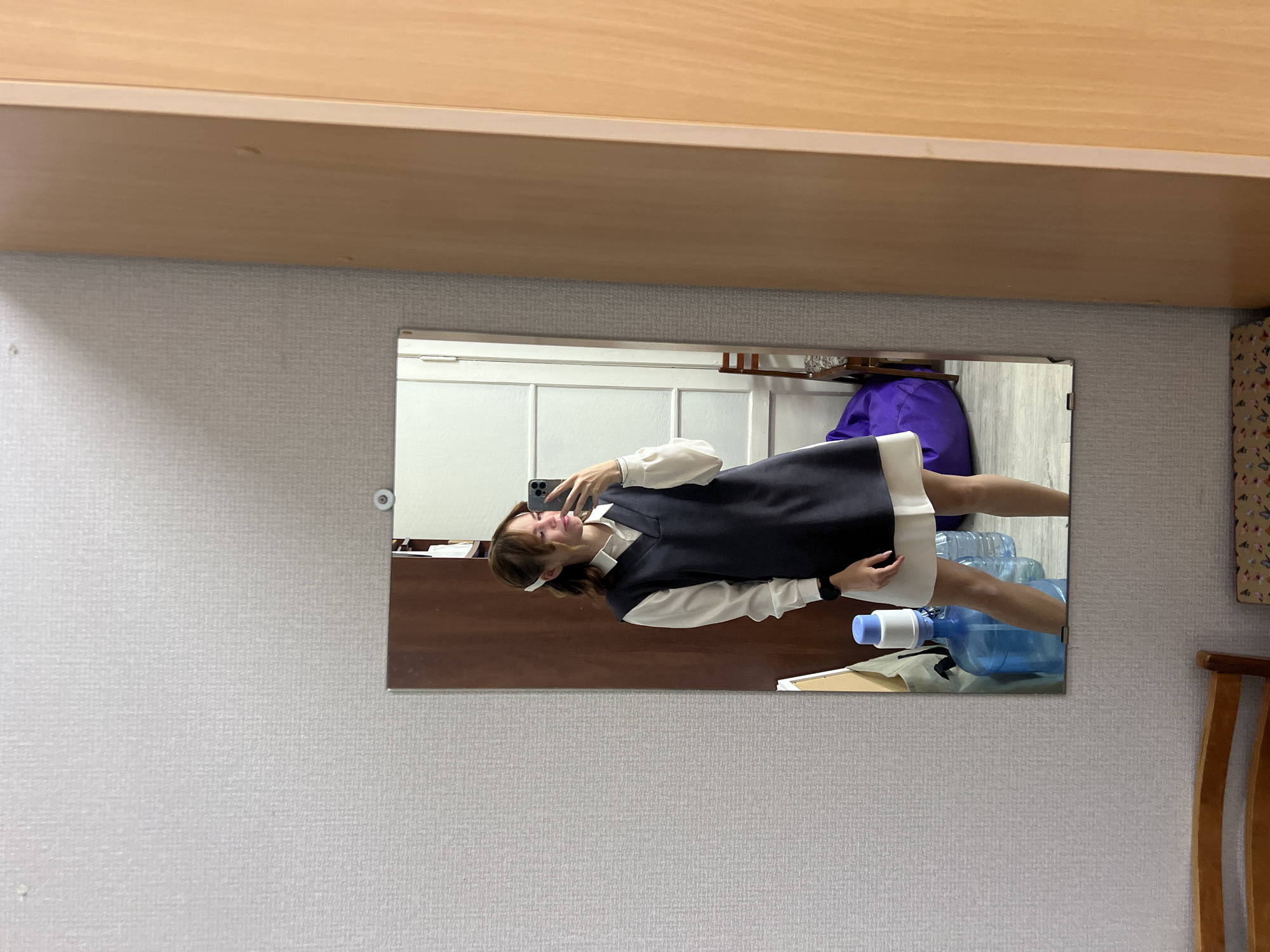 A young woman is taking a mirror selfie in an indoor setting. She is wearing a white long-sleeved shirt and a dark V-neck pinafore dress. Her hair is pulled back with a white hair accessory. The mirror reflects a portion of the room, showing large water cooler bottles and a purple beanbag chair. The room appears to be a residential space, likely an apartment, possibly located in Chaikovsky, Russia, as indicated by the context. The wall texture and the style of the wardrobe suggest a common interior design found in Russian apartments.